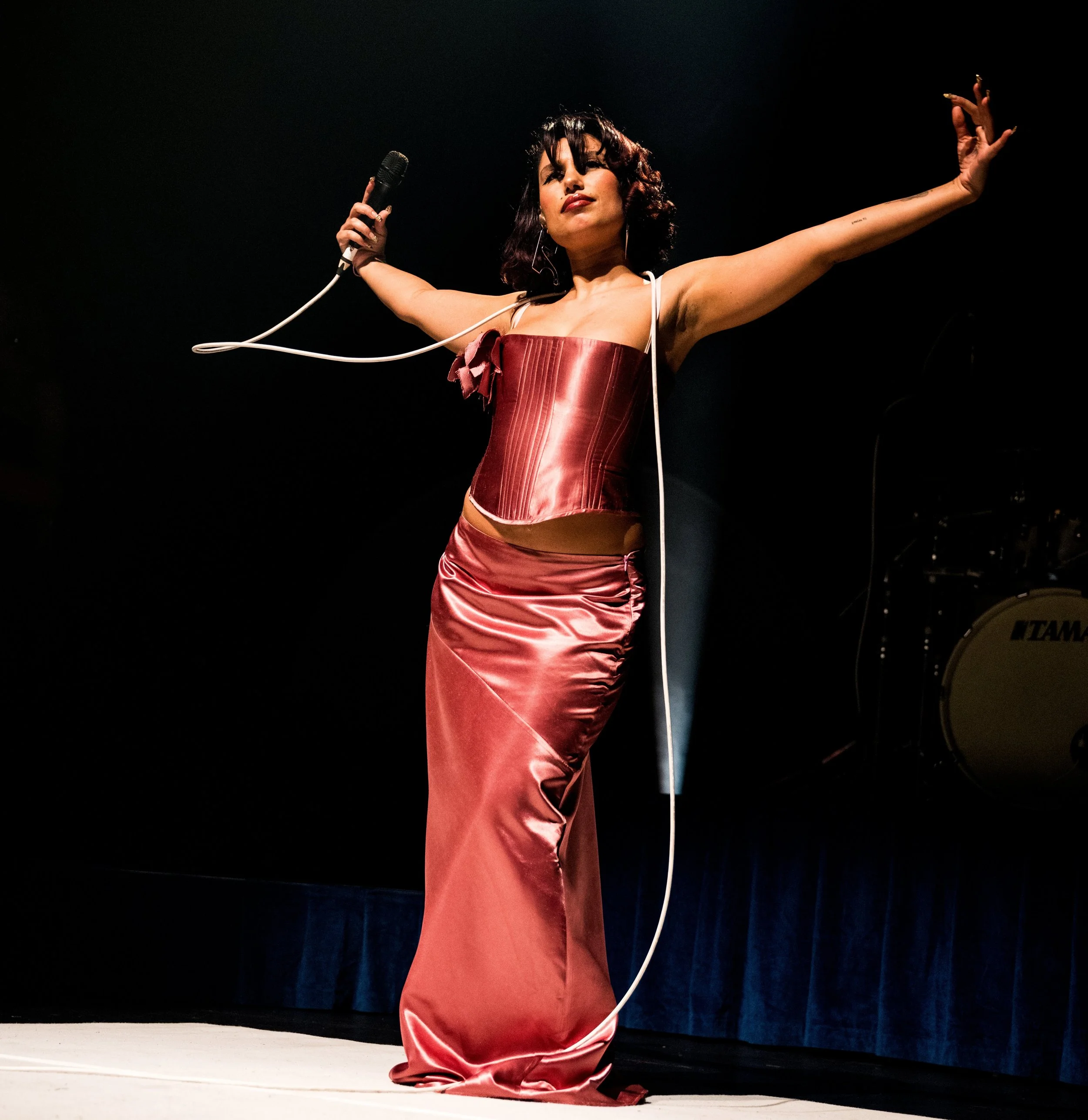 A woman in a shiny red satin dress and matching corset performing on stage with her arms outstretched, holding a microphone in one hand.