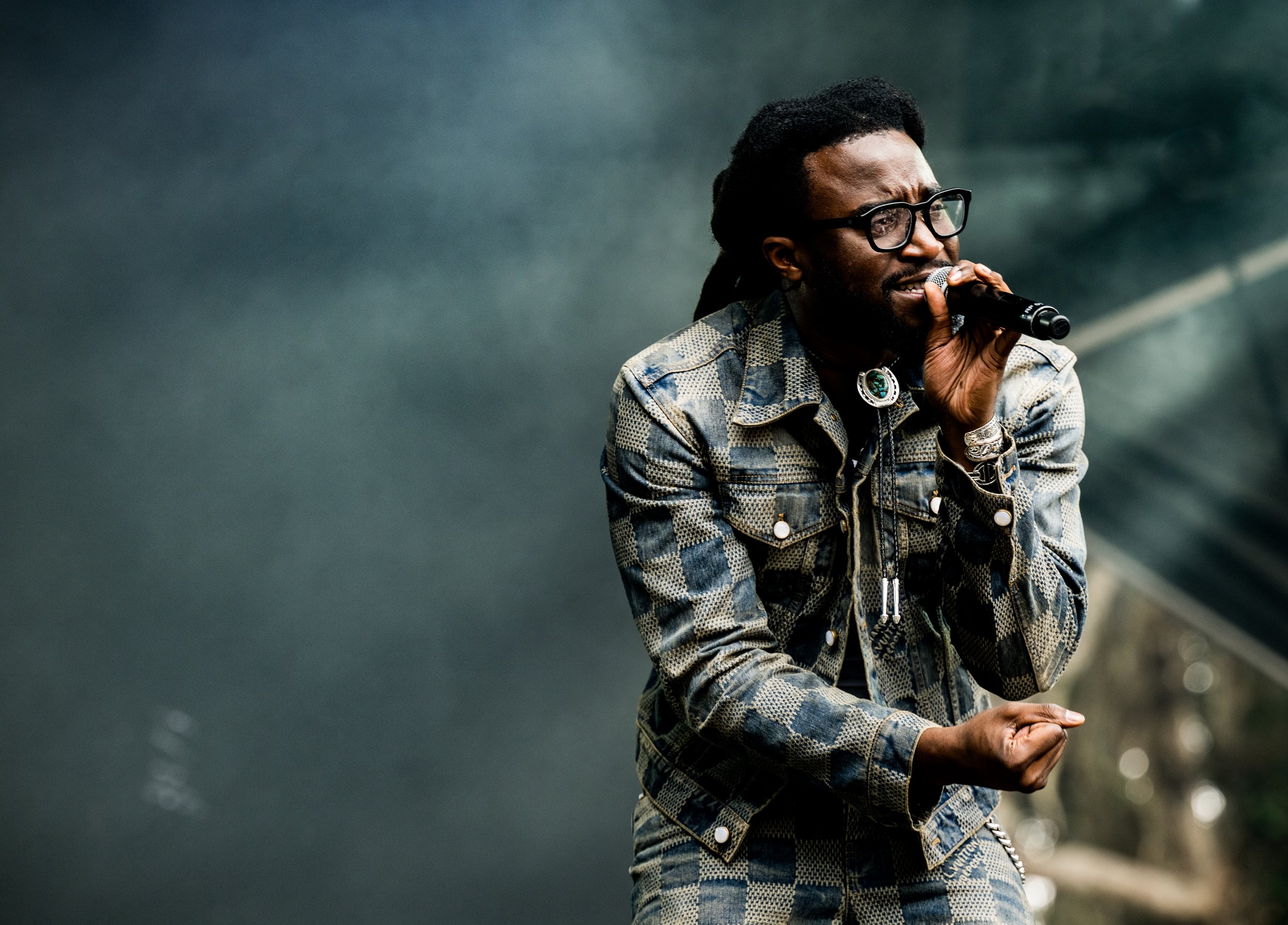 A man with dreadlocks and glasses holding a microphone, performing on stage with a dark background.