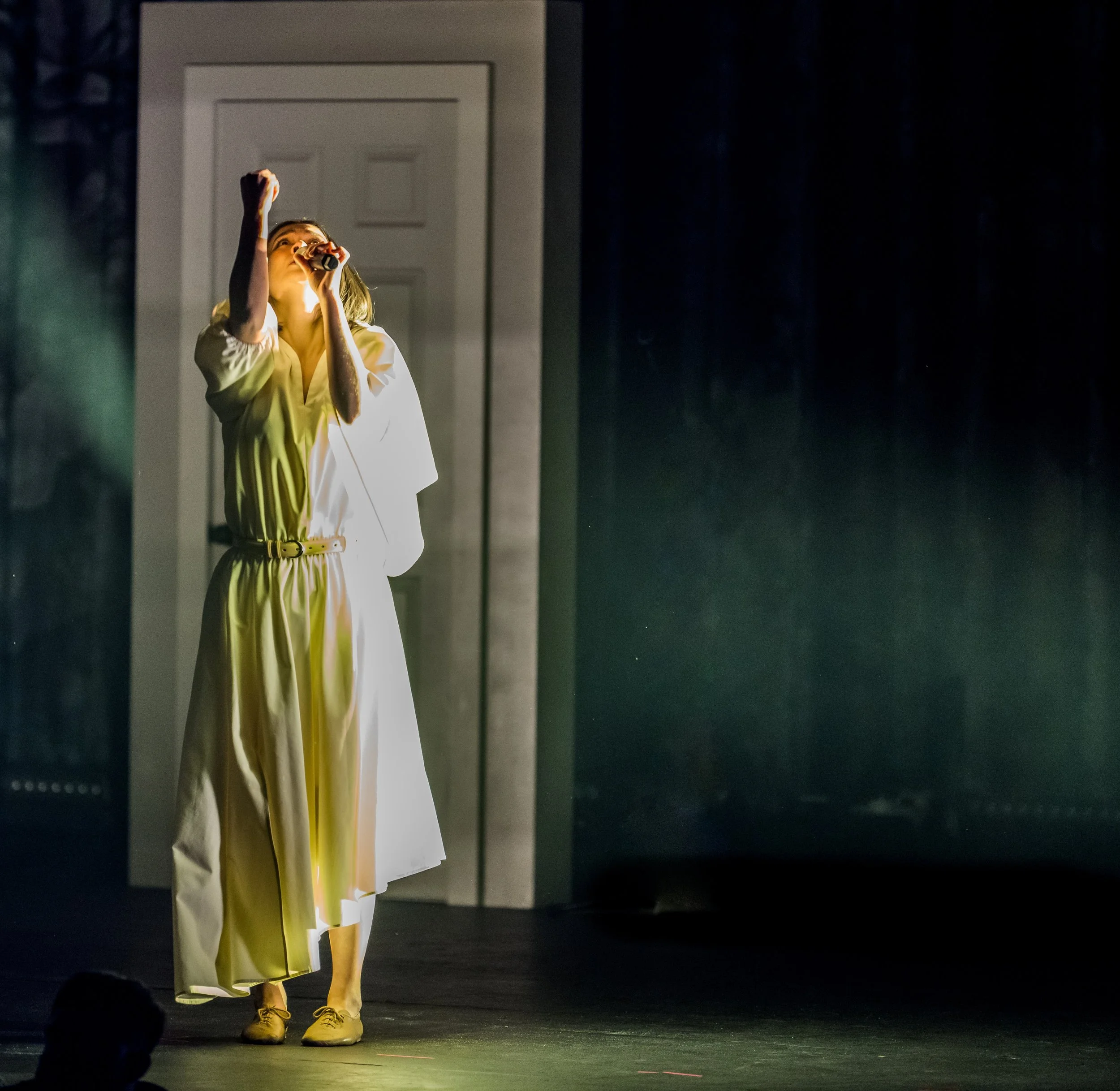 A woman in a white dress performs on stage, holding a microphone, with a door in the background and stage lighting highlighting her.