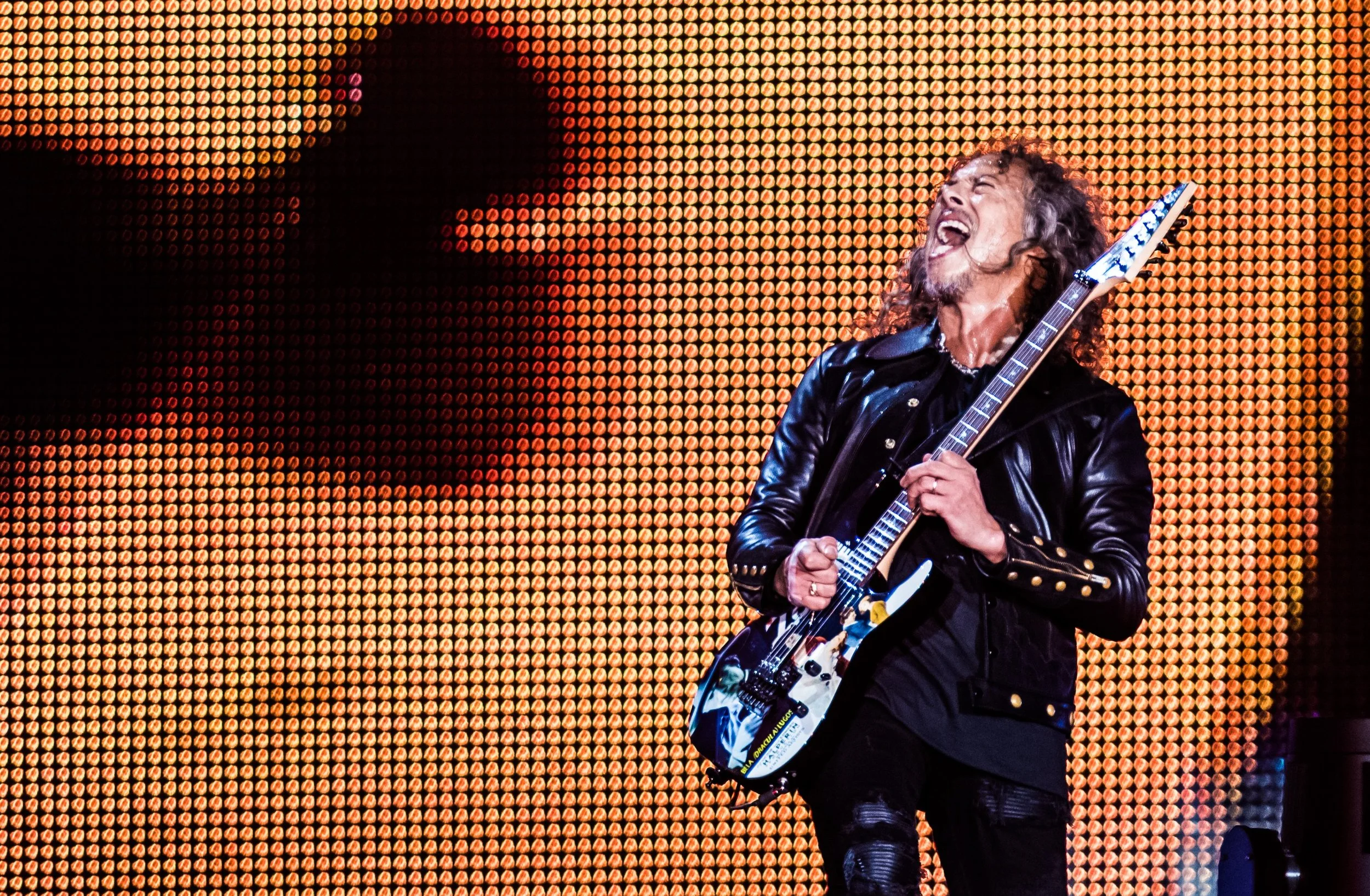 A male guitarist passionately performing on stage with a vivid orange and yellow LED background.