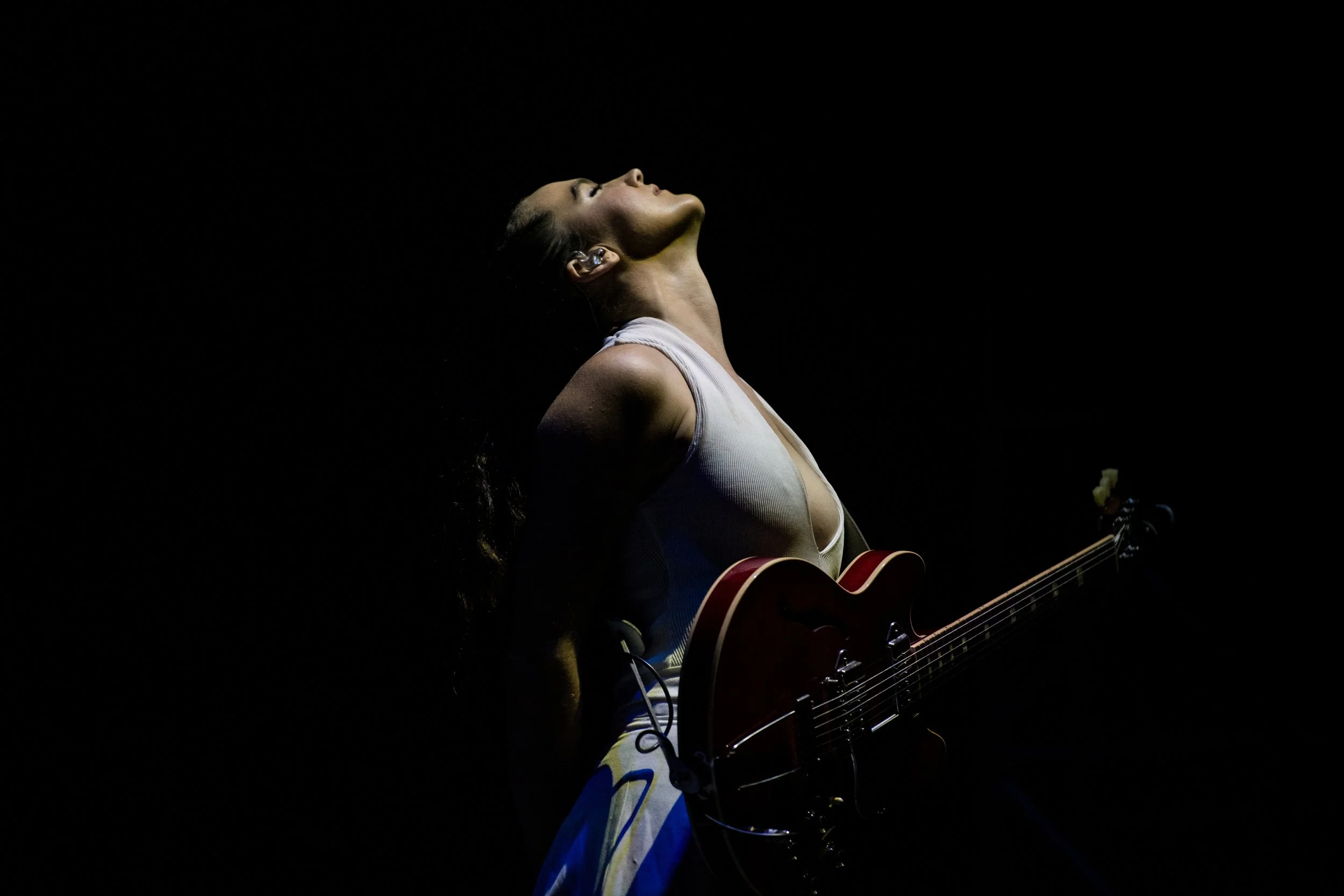 A woman performing on stage with a guitar, illuminated in a dark background, looking upwards with her eyes closed.