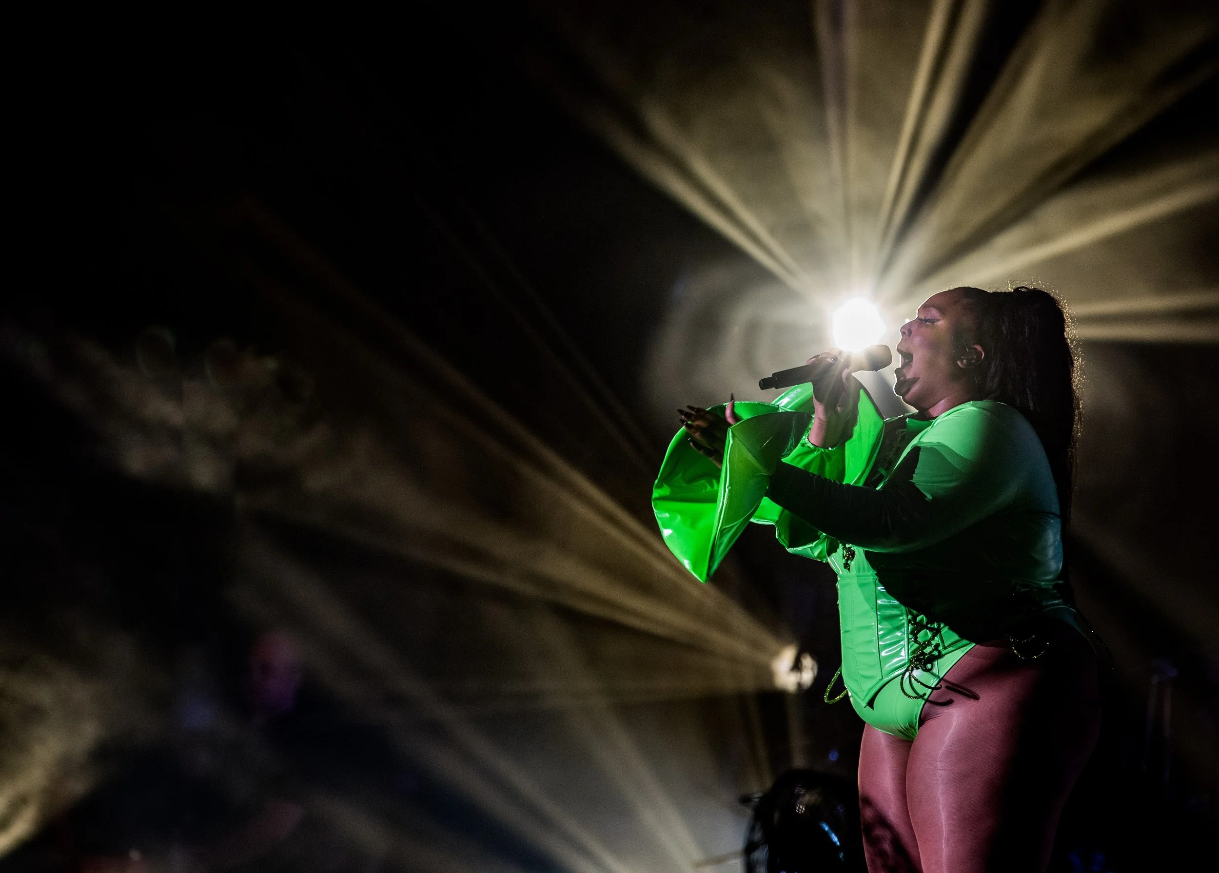 A female singer performing on stage, wearing a green outfit with the background illuminated by bright lights.