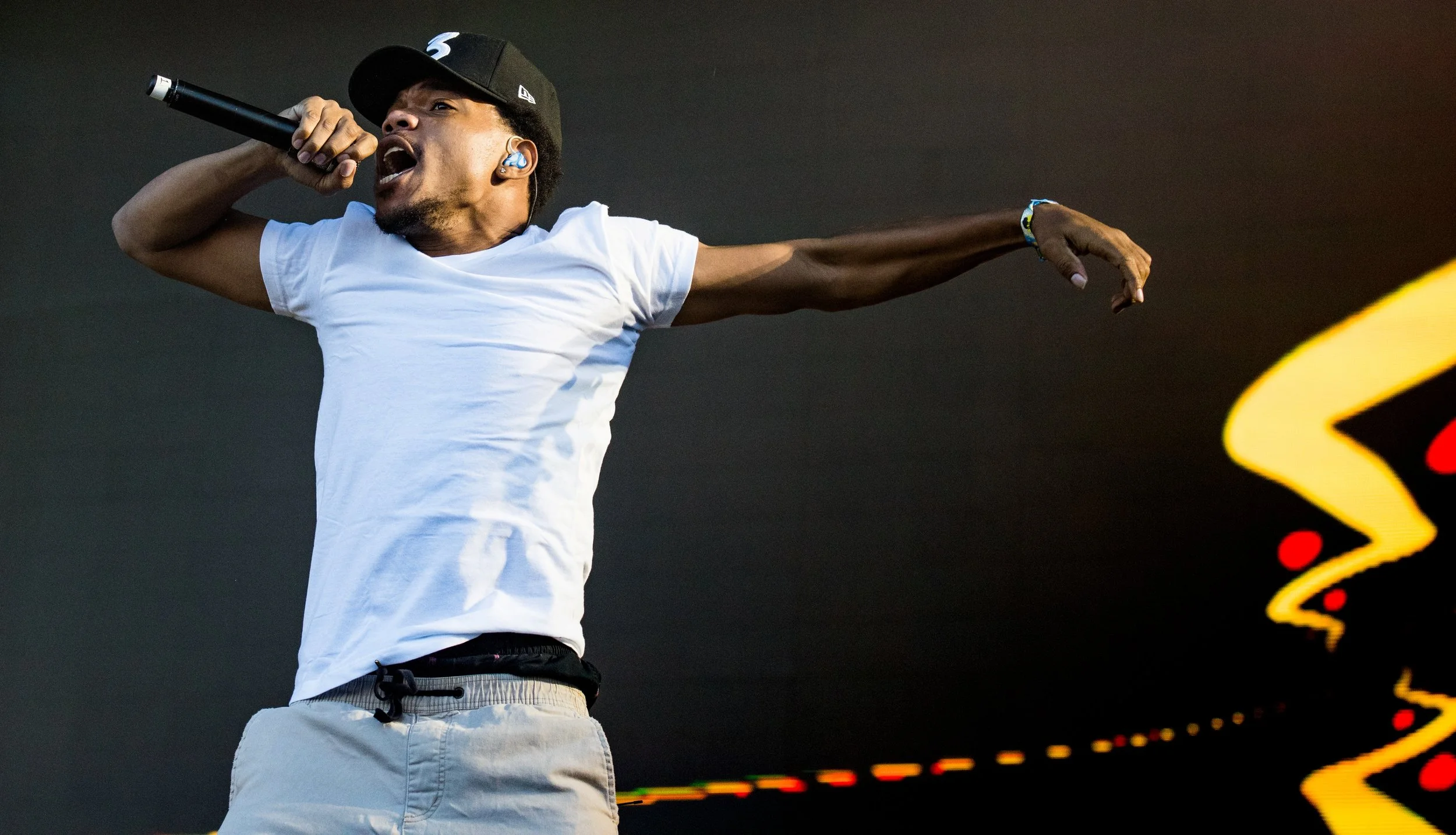 A male performer singing into a microphone on stage, wearing a white t-shirt, beige shorts, a black cap, and blue in-ear monitors, with his left arm extended outward.