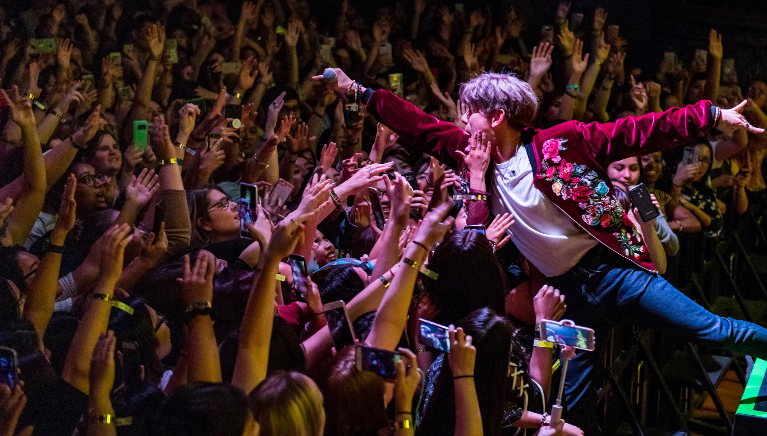 Performer reaching out to an enthusiastic crowd at a concert or event, with many audience members holding up phones and wearing yellow wristbands.