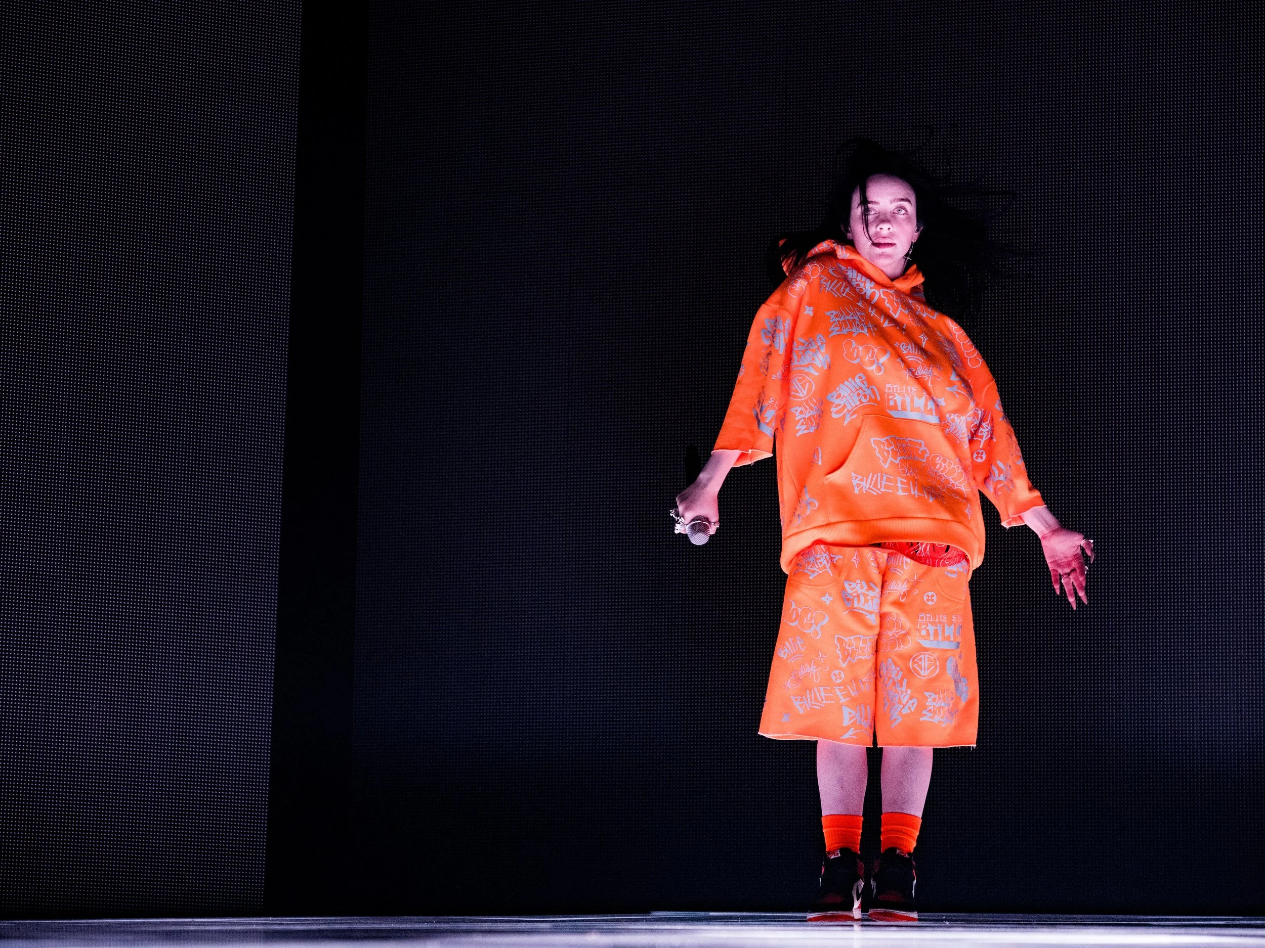 A person standing on stage wearing an orange outfit with graffiti-style writing, holding a microphone in one hand, and appearing surprised or shocked.