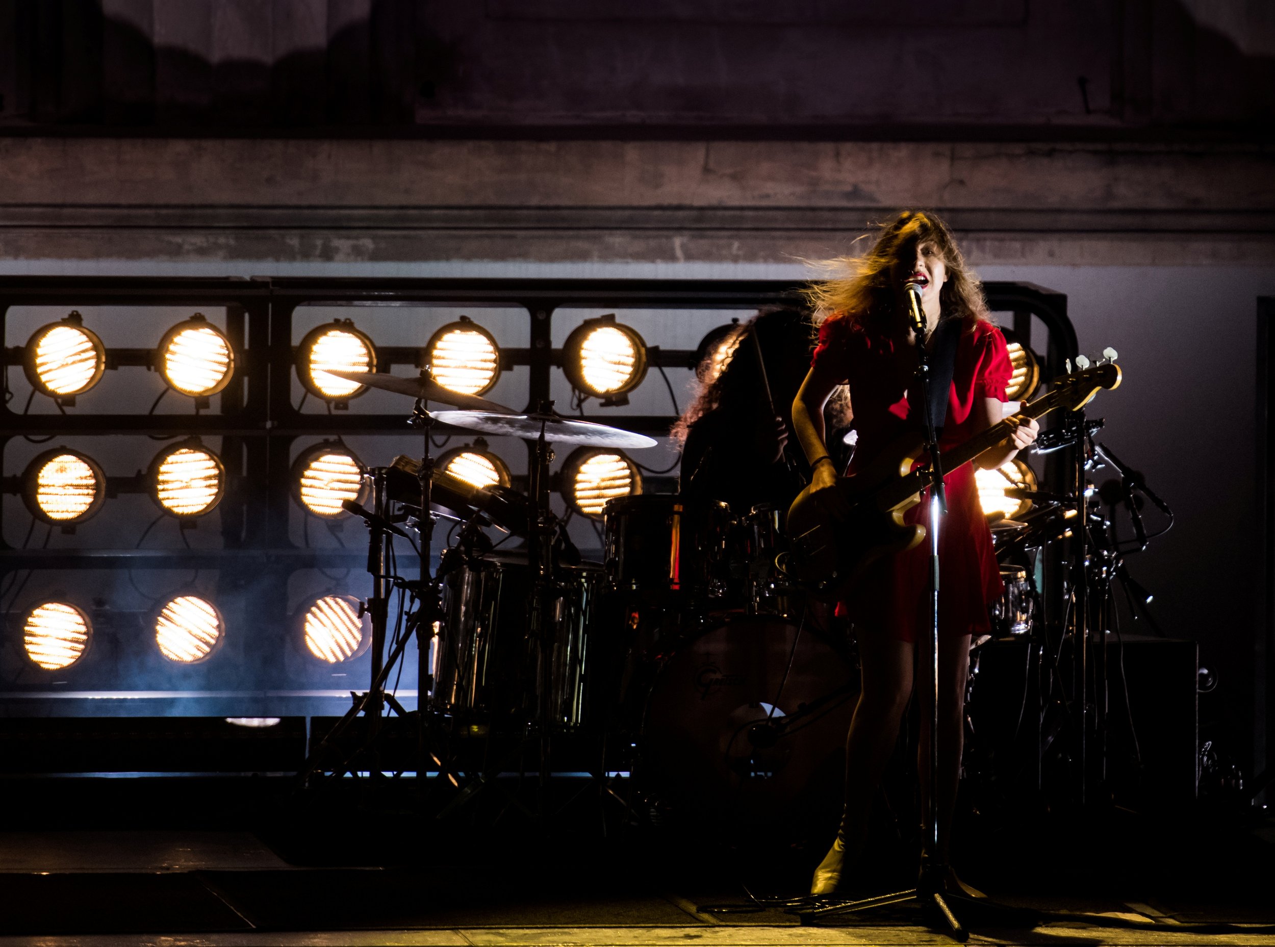 A female singer wearing a red dress plays a guitar on stage with bright yellow stage lights behind her.