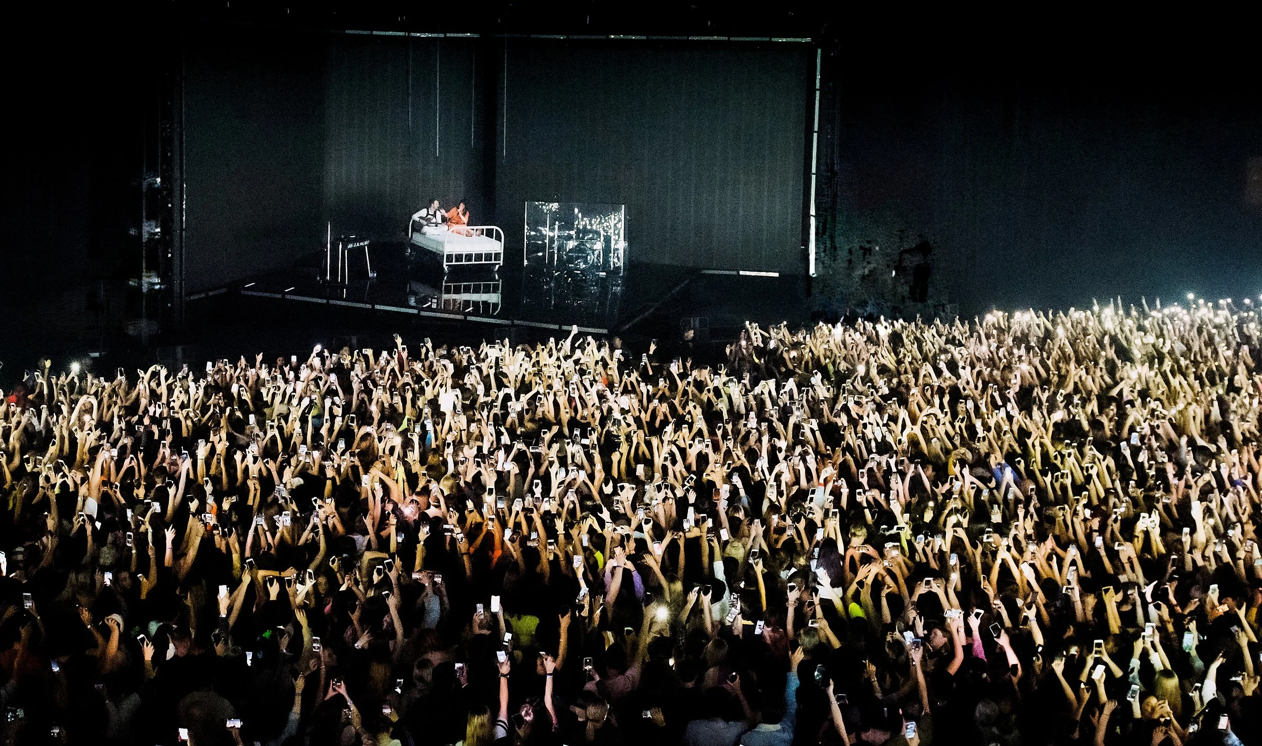 Crowd at concert with many people holding smartphones, stage in the background with performers, dark setting, and focused lighting on stage.