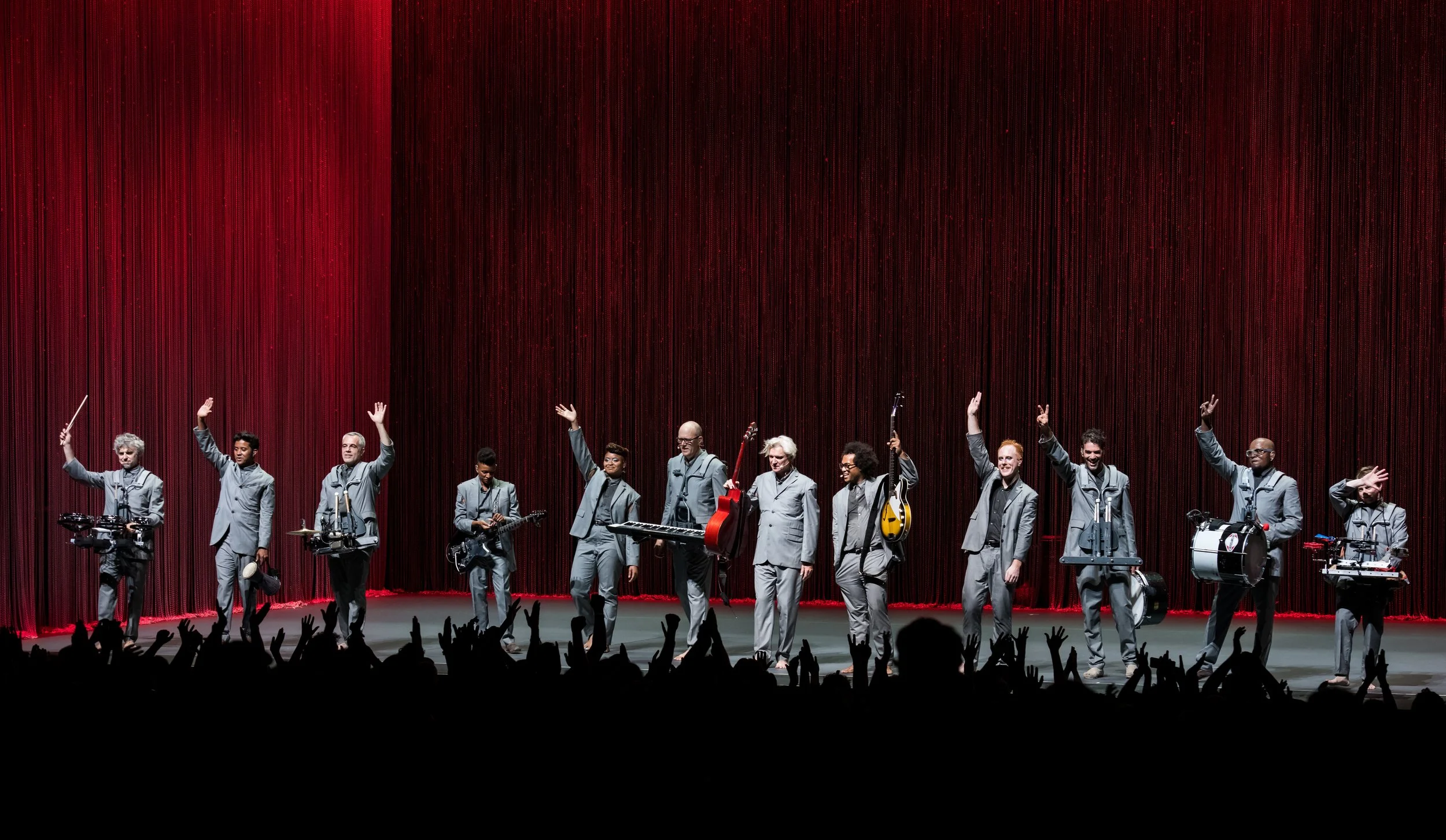 A group of twelve musicians on stage, each holding musical instruments, bowing and waving to an audience, in front of a red curtain.