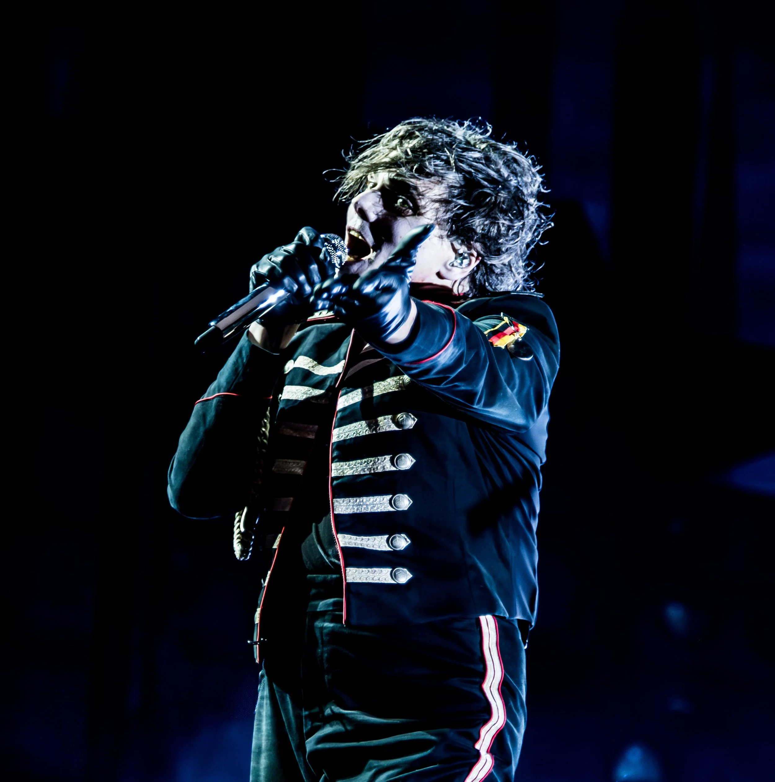 A performer in a military-style jacket with gold buttons and decorations is singing or speaking into a microphone on stage, with expressive gestures.