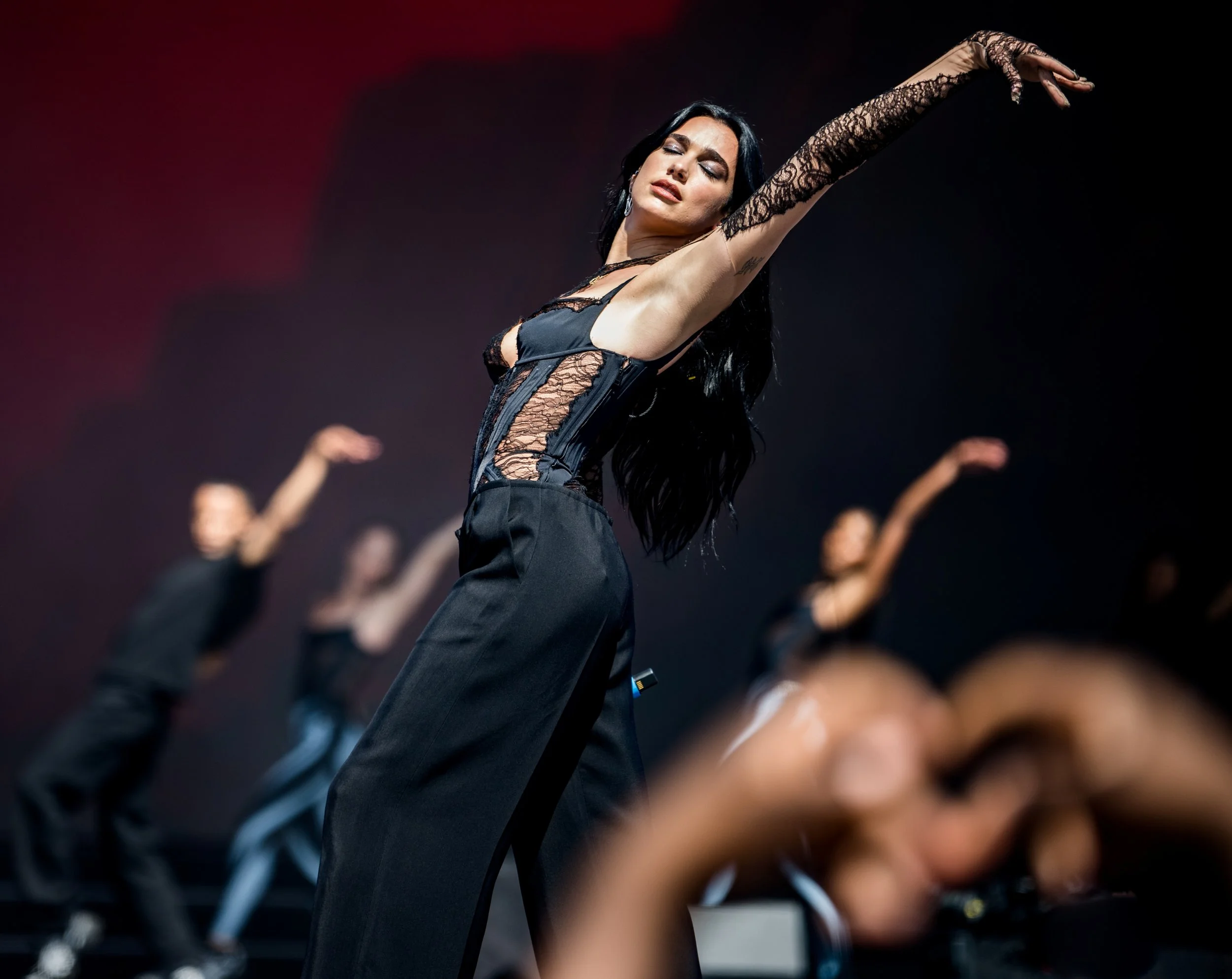 A woman with long dark hair in a black lace top and black pants performs a dance move with her arm raised and eyes closed on stage, with other dancers blurred in the background.