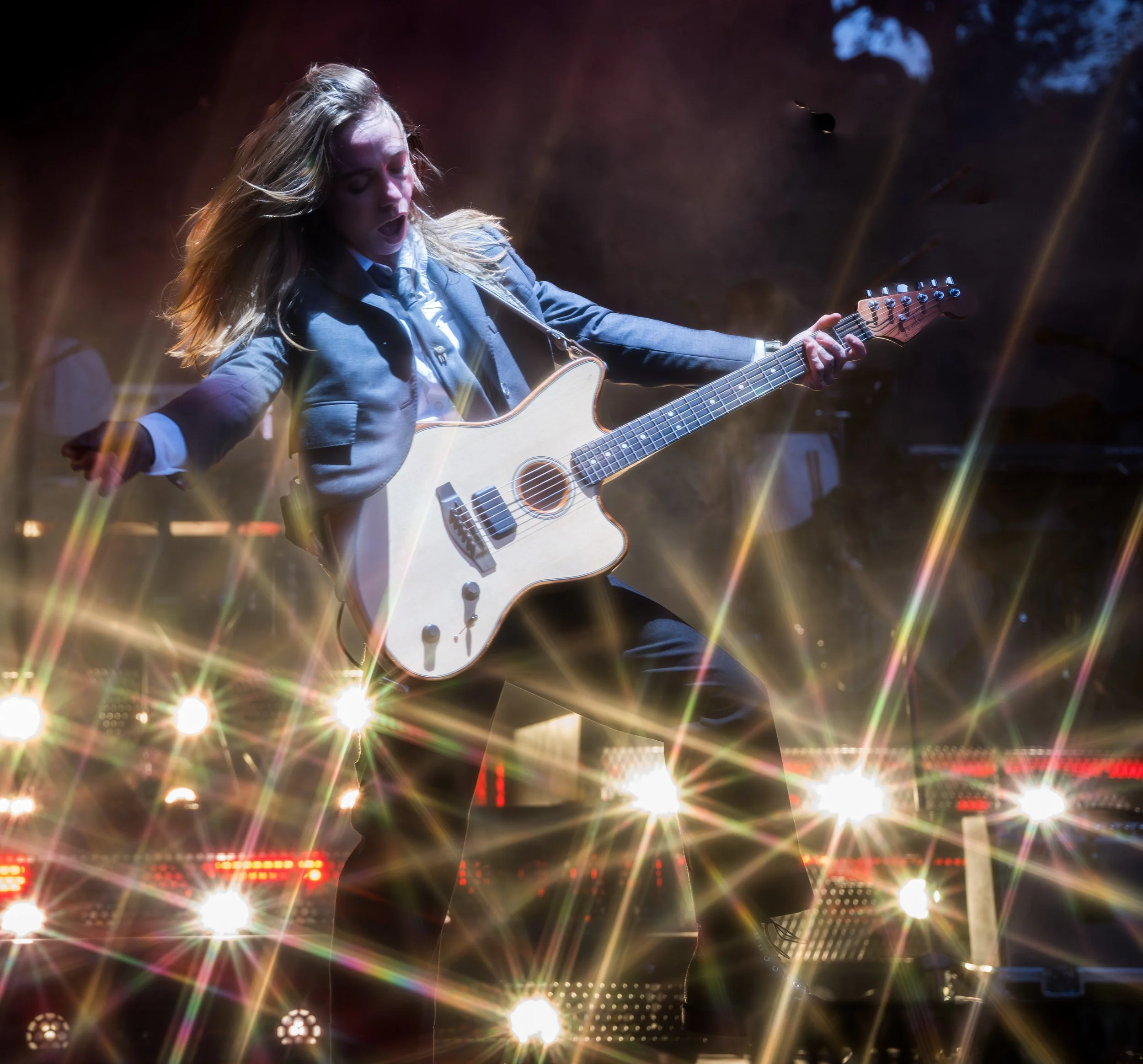 A female musician plays an electric guitar on stage at night, surrounded by bright stage lights creating a starburst effect.