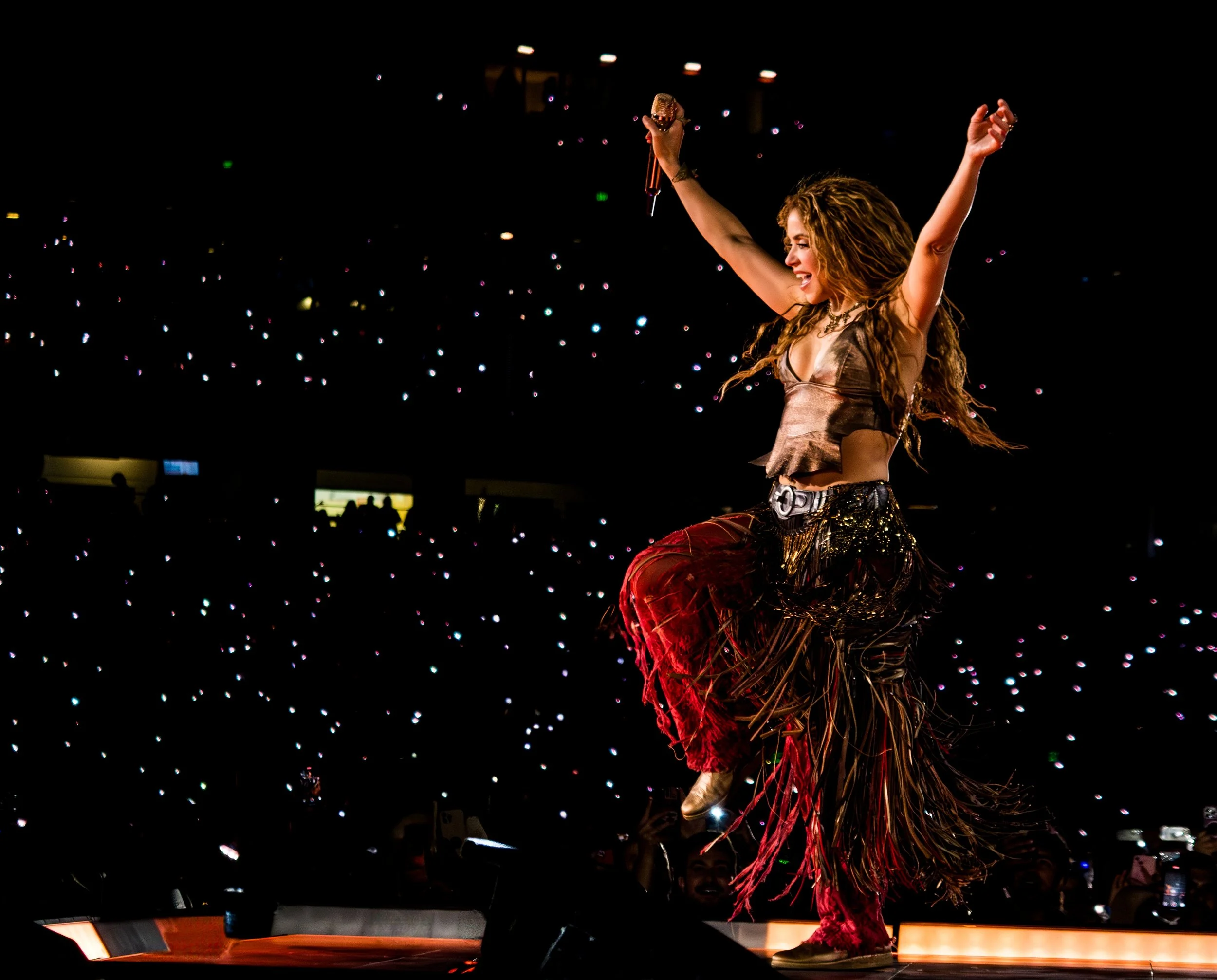 A woman performing on stage with her arms raised, holding a microphone, in a brightly lit arena filled with lights and audience.
