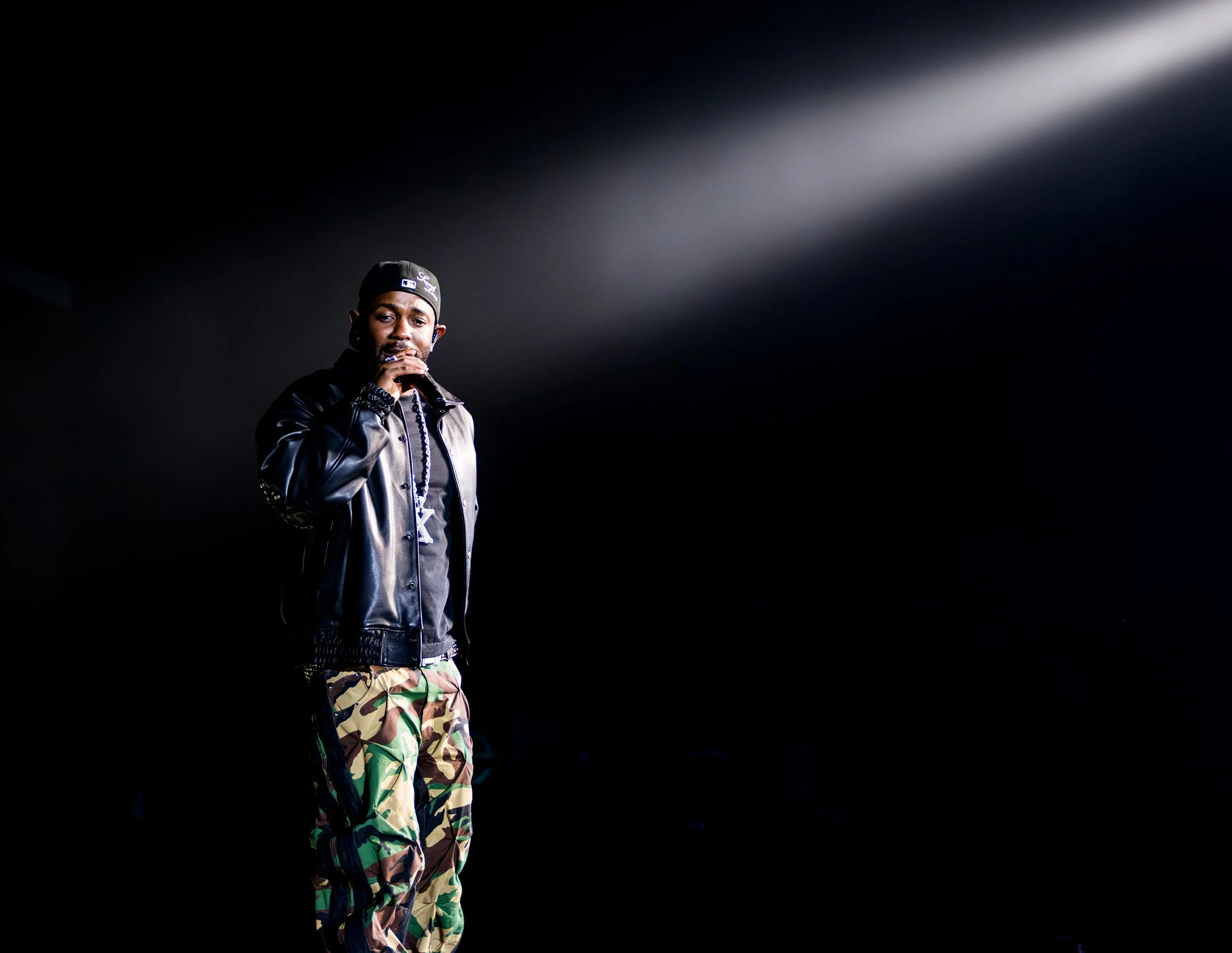 A male performer stands on stage holding a microphone, dressed in a black leather jacket, camouflage pants, wearing a black cap, with a serious expression, against a dark background with a spotlight shining on him.