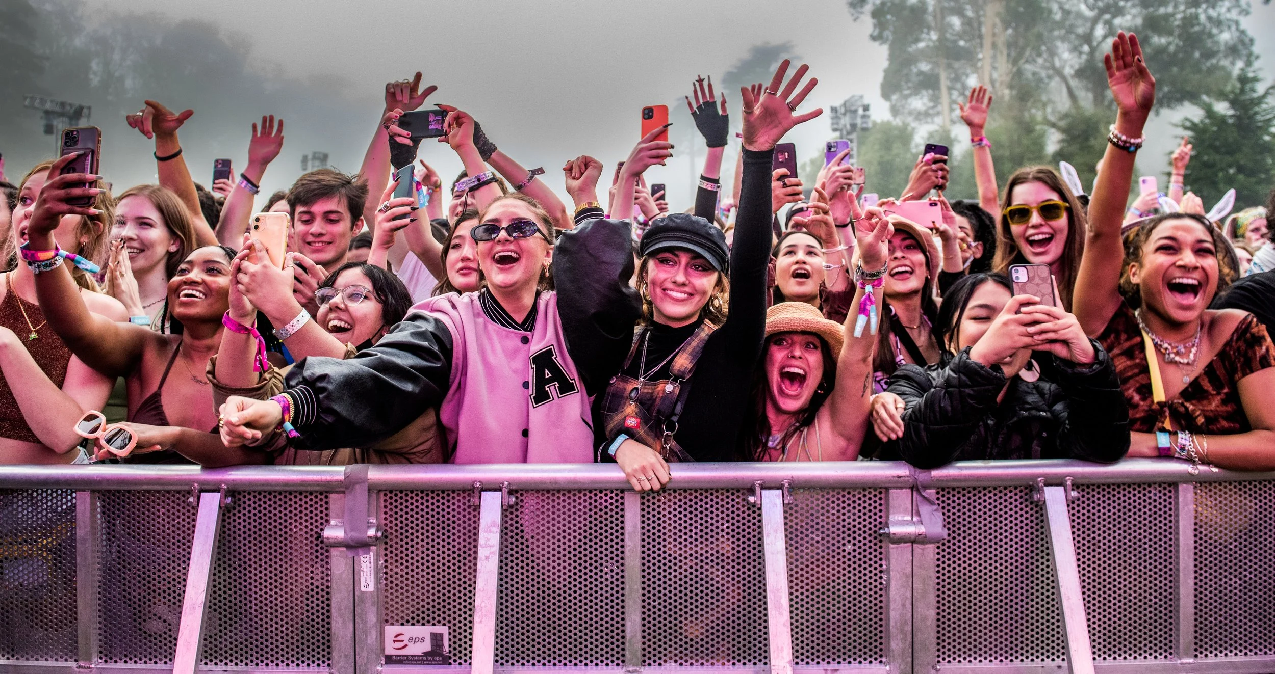 An excited crowd of young concert-goers standing behind a metal barricade, cheering, smiling, and taking photos at an outdoor music event.