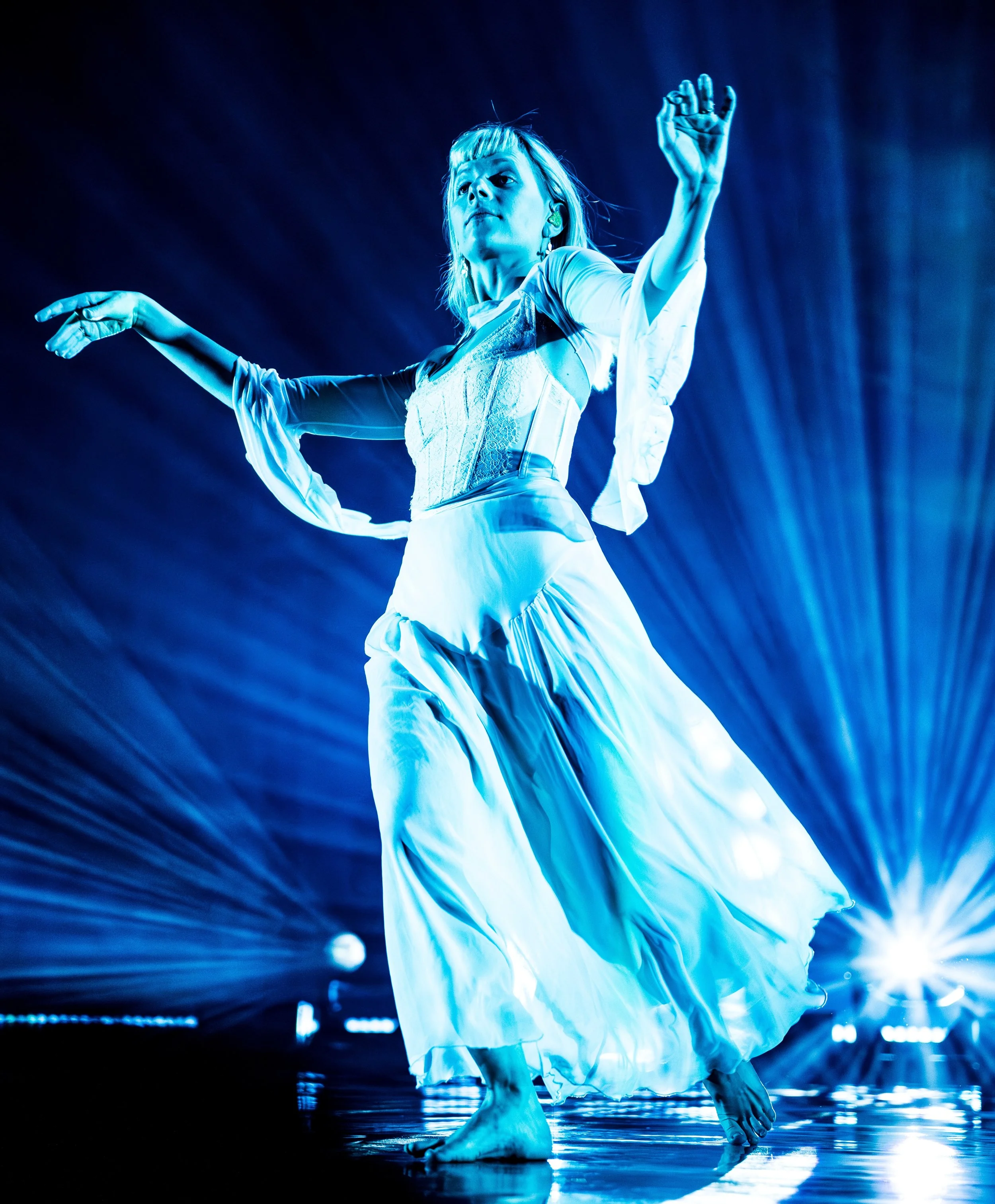 A woman dancing barefoot on stage, illuminated by vibrant blue stage lights, wearing a flowing white skirt and a fitted white top, with her arms raised gracefully.
