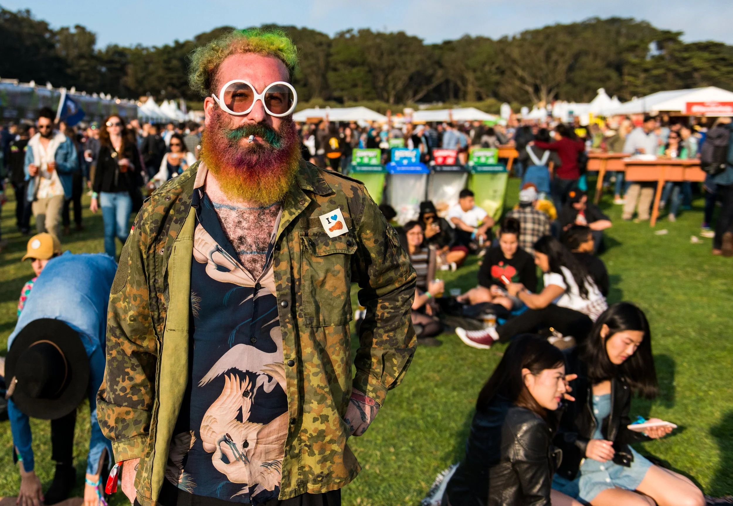 A man with a rainbow-colored beard and hair wearing large white sunglasses, camouflage jacket, and a colorful shirt with a dragon design, standing outdoors at a crowded festival or park.
