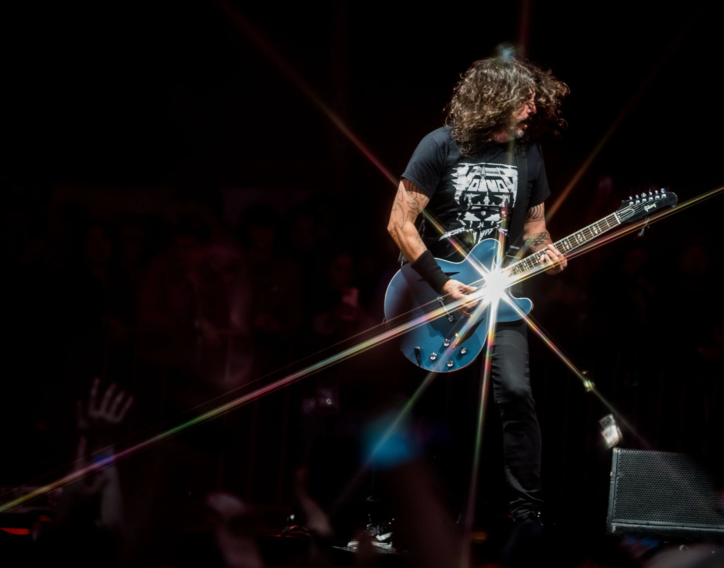 A musician with long curly hair playing an electric guitar on stage with dramatic lighting and lens flare effect.