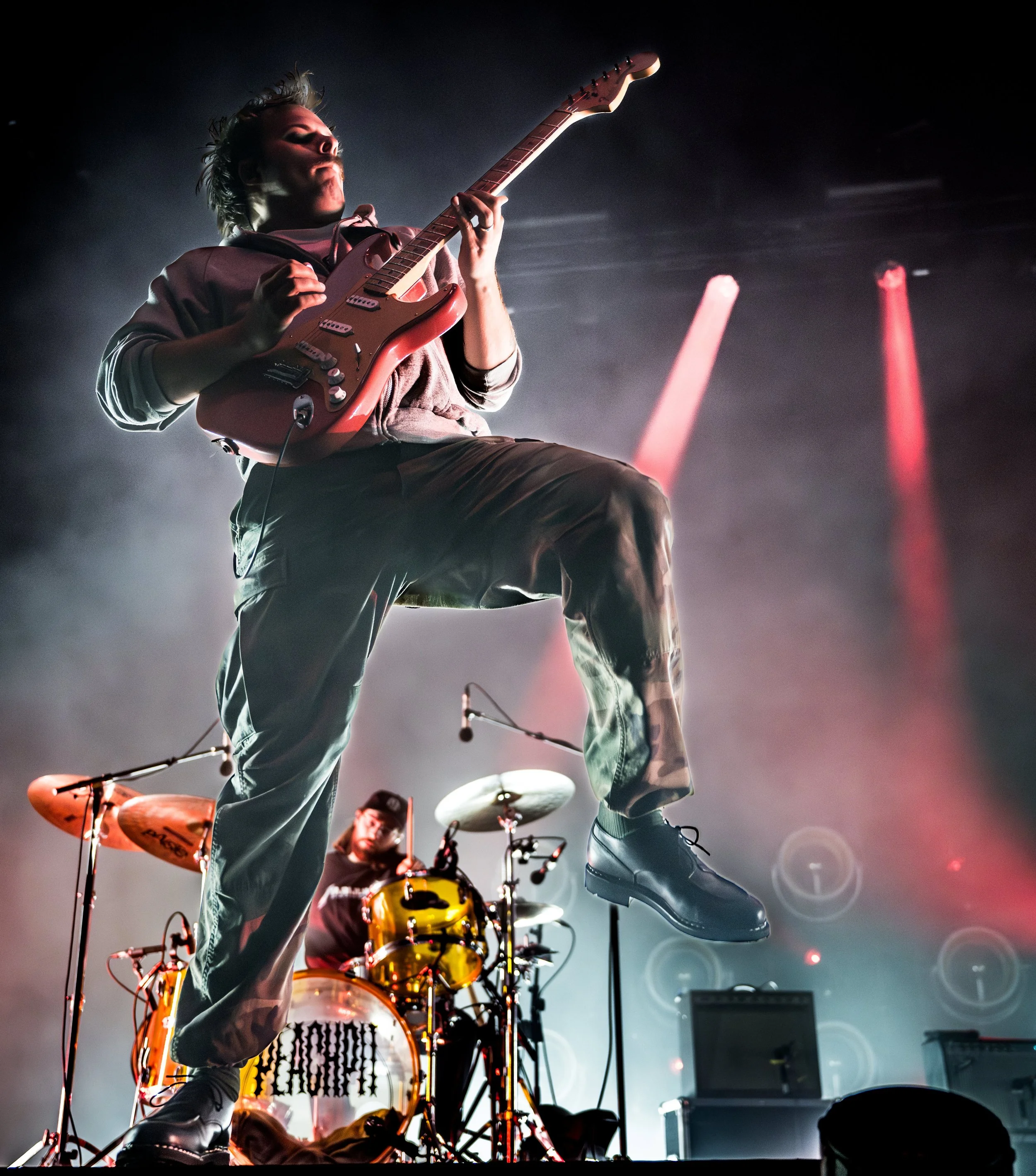 Musician playing an electric guitar during a live performance, with a drummer in the background, stage lights, and fog creating a concert atmosphere.