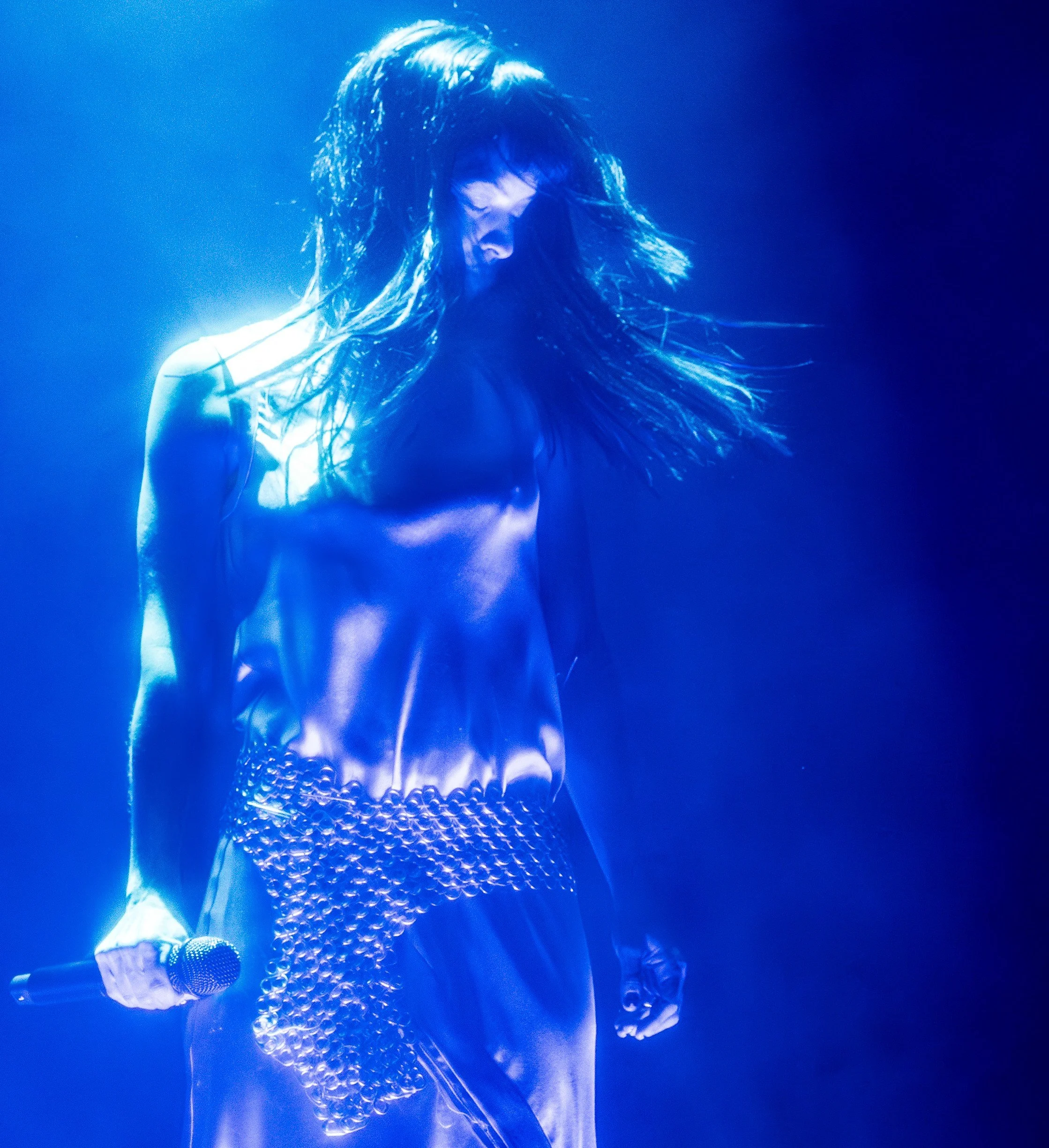 Female performer with long hair, shirtless, holding a microphone, illuminated by blue stage lighting.