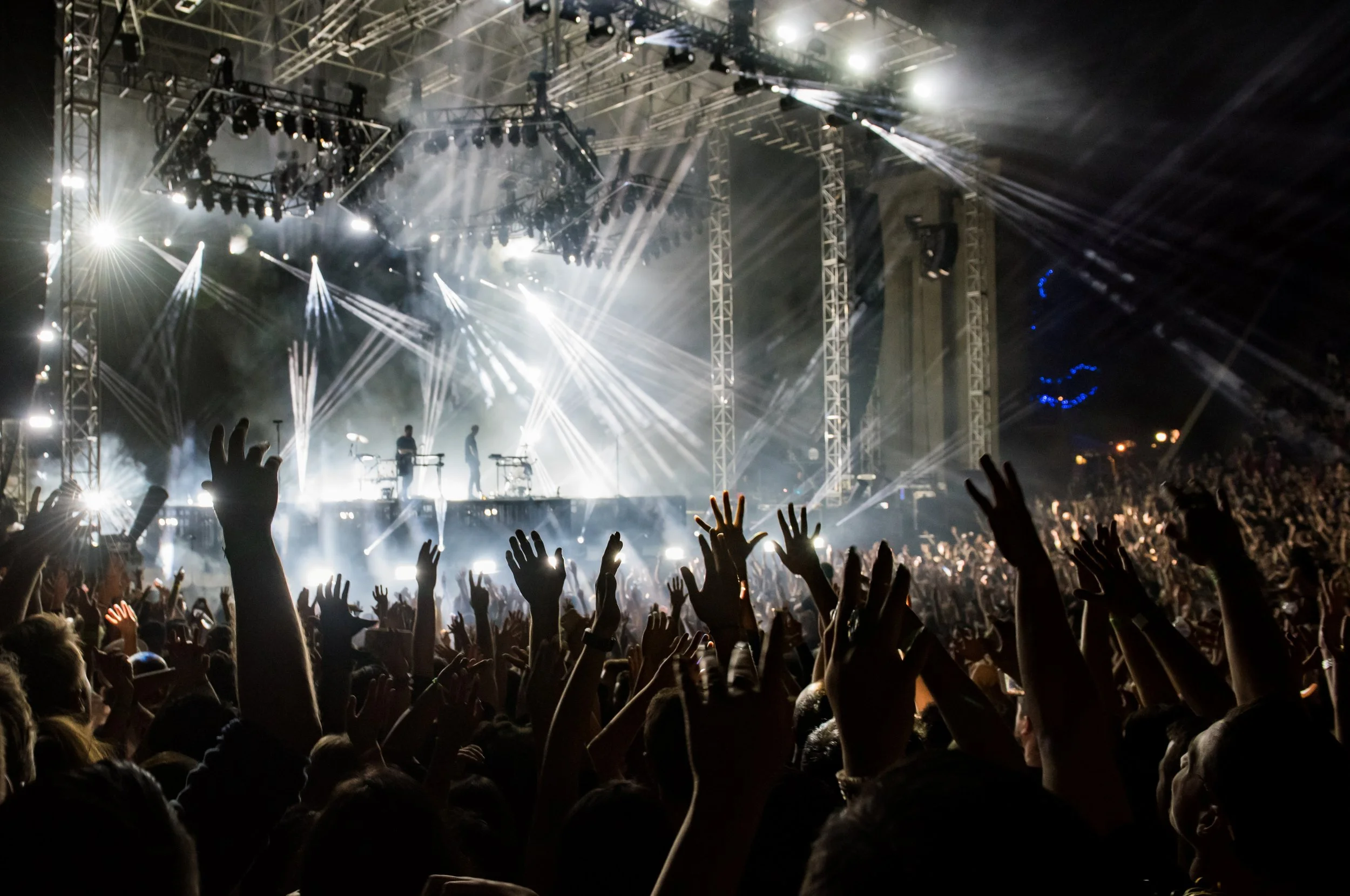 Crowd at a concert with raised hands facing a brightly lit stage with performers and stage lighting effects.