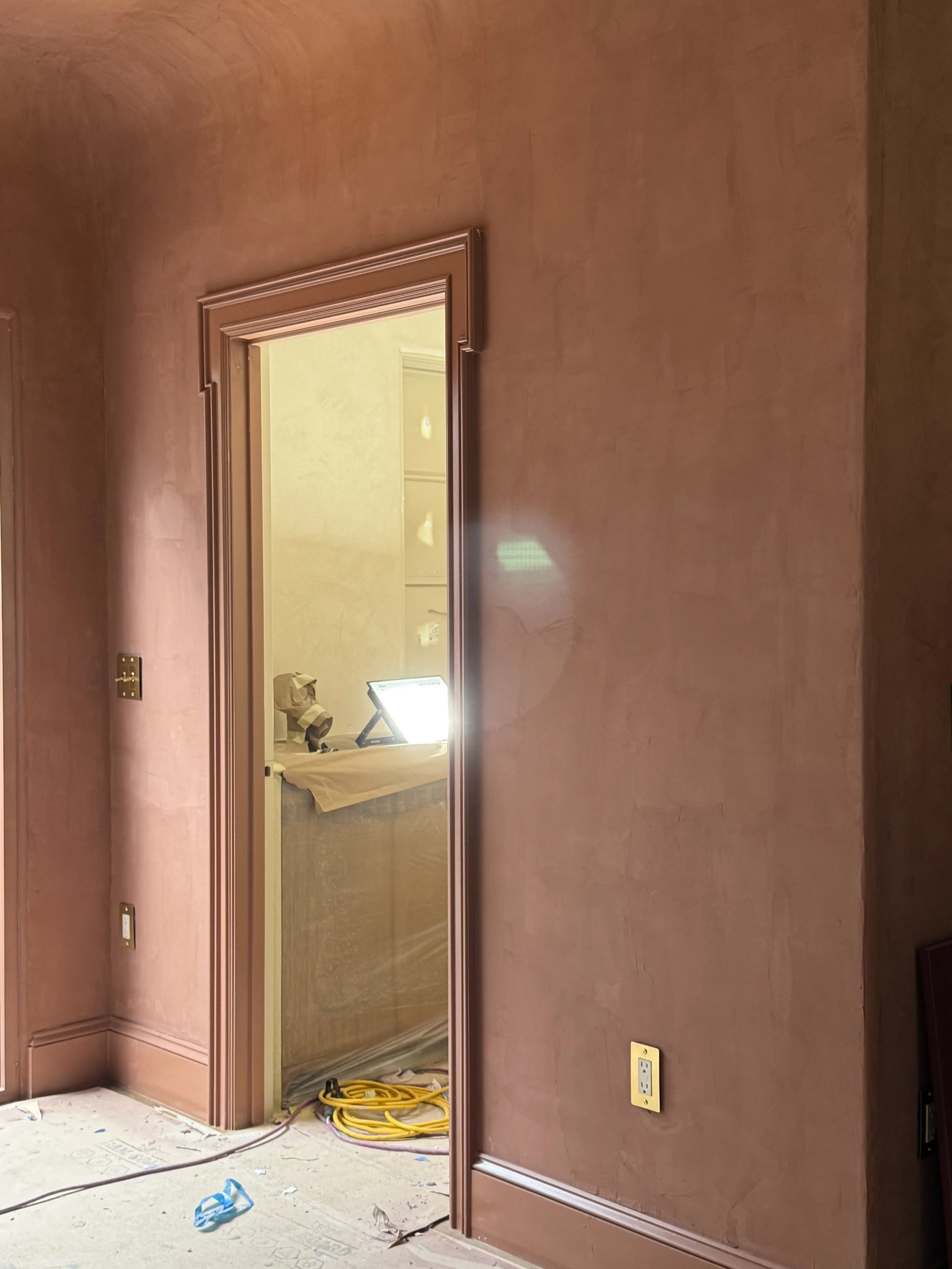 A partially visible room under construction with brown walls and a doorway leading to a lit room with a table and tools.