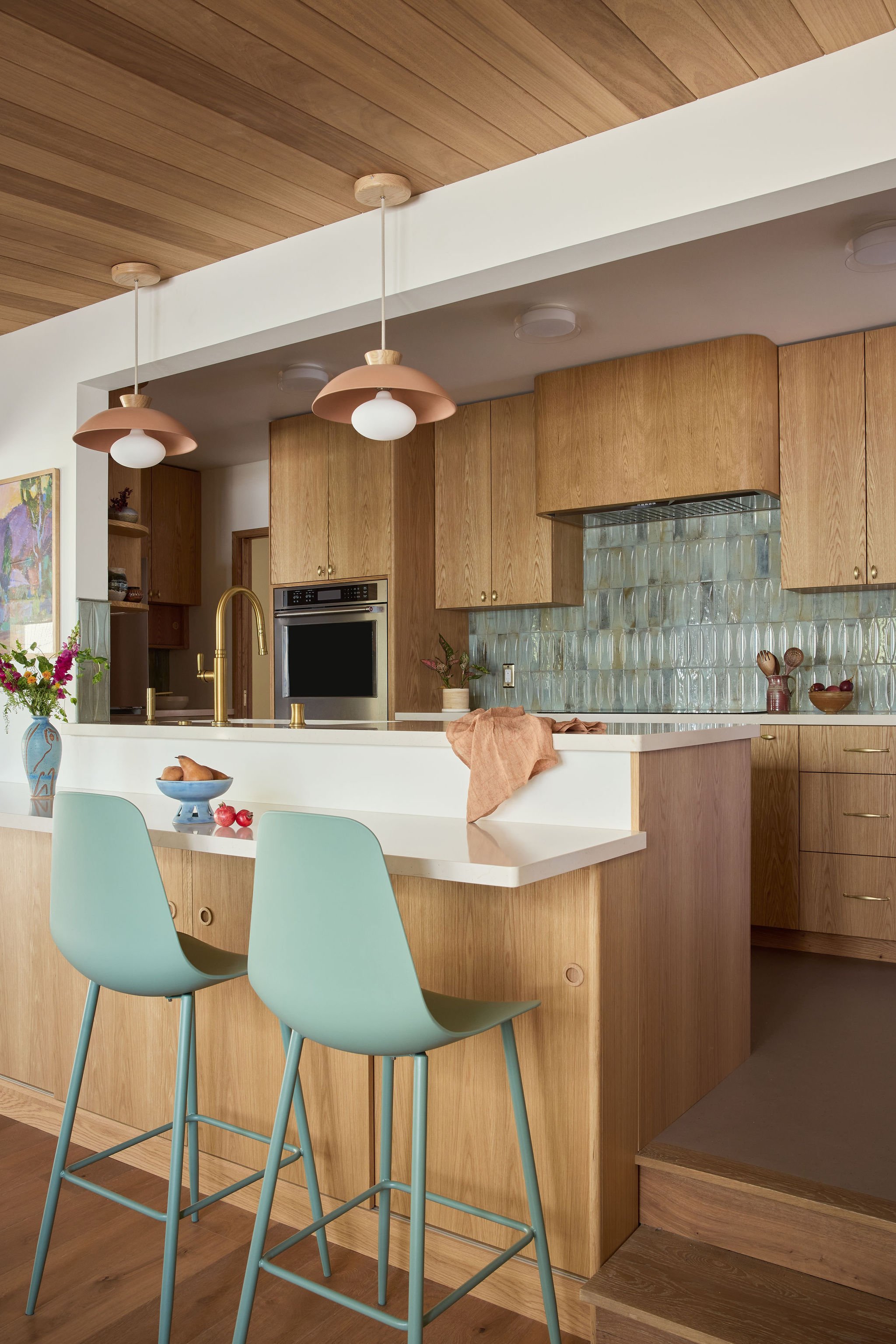 Kitchen with wooden cabinets, a glass backsplash, and two mint green bar stools at a white countertop. Pendant lights with pink shades hang from the wooden ceiling.