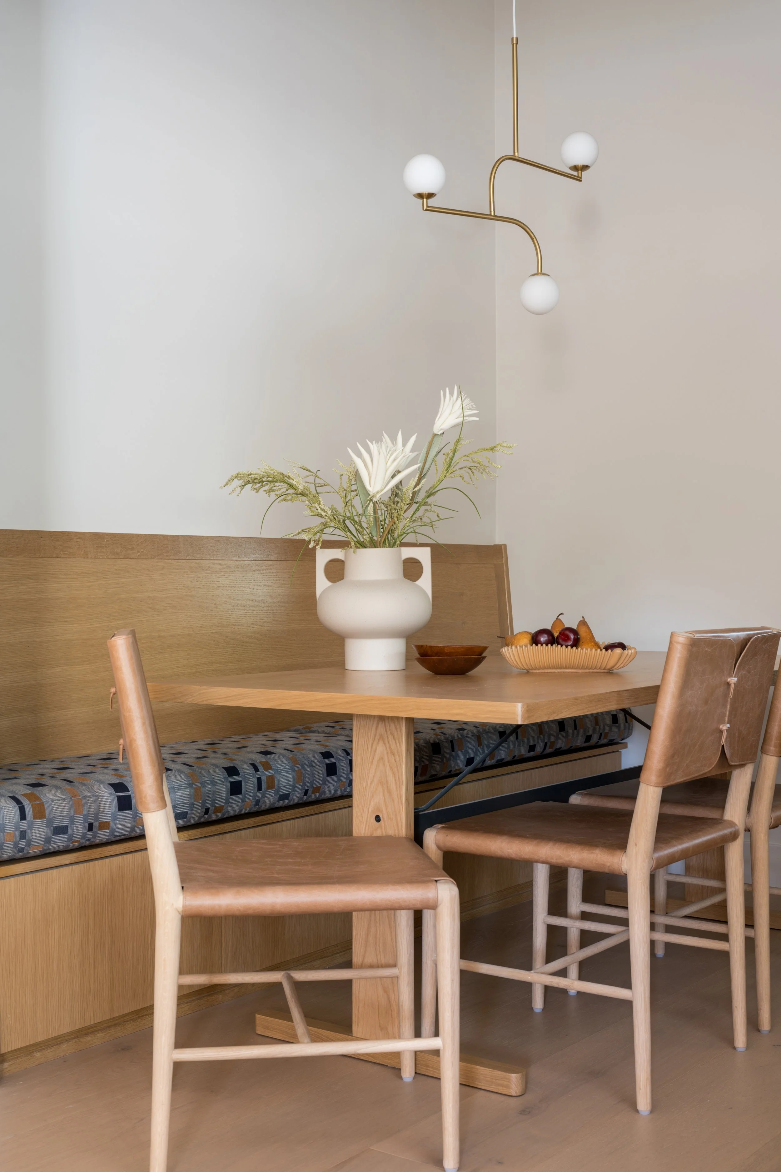 A dining area with a wooden table, two wooden chairs, a built-in wooden bench with a patterned cushion, a white vase with flowers, a small wooden bowl, and a fruit bowl with assorted fruits. A modern ceiling light fixture hangs above.
