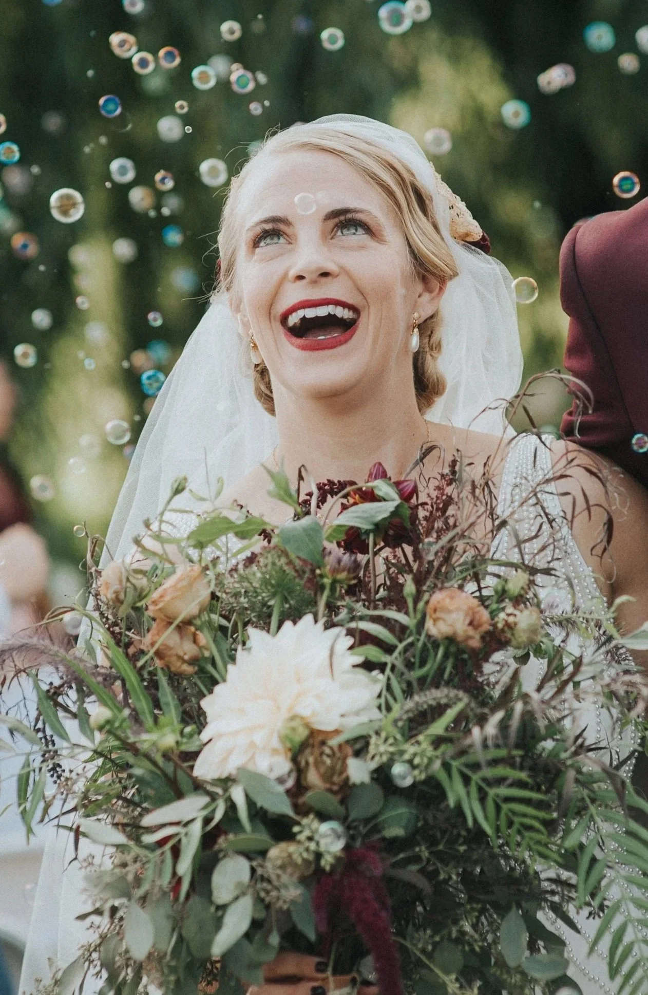 A smiling bride in a wedding dress holding a large bouquet of flowers, surrounded by hanging bubbles outdoors.