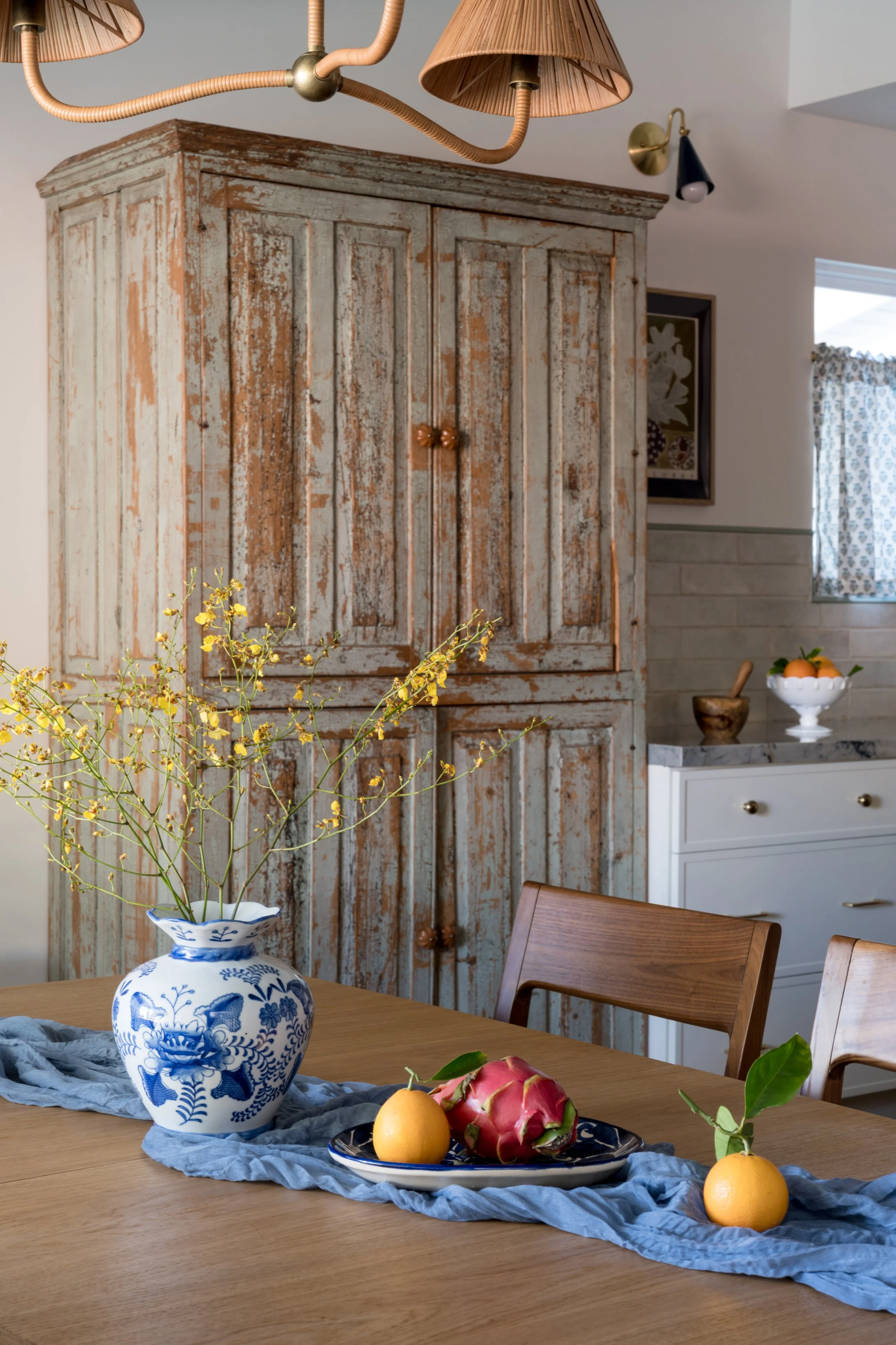 A rustic wooden cabinet with weathered paint, a dining table with a blue cloth runner, a blue and white floral vase with yellow flowers, a plate with fruit (lemons and a dragon fruit), and a wooden chair. In the background, there is a white sideboard with a bowl of oranges, a wooden mortar and pestle, and a window with patterned curtains.
