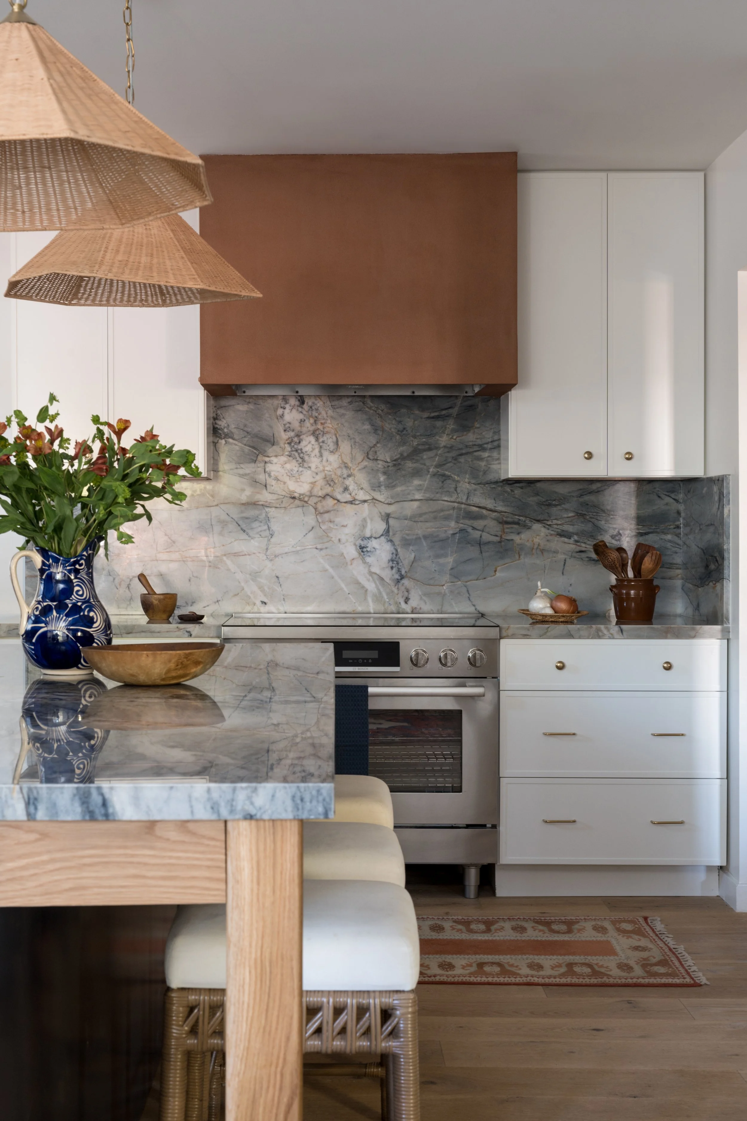 Kitchen with marble backsplash and counter, white cabinets, stainless steel oven, and decorative items including flowers, bowls, and utensils.