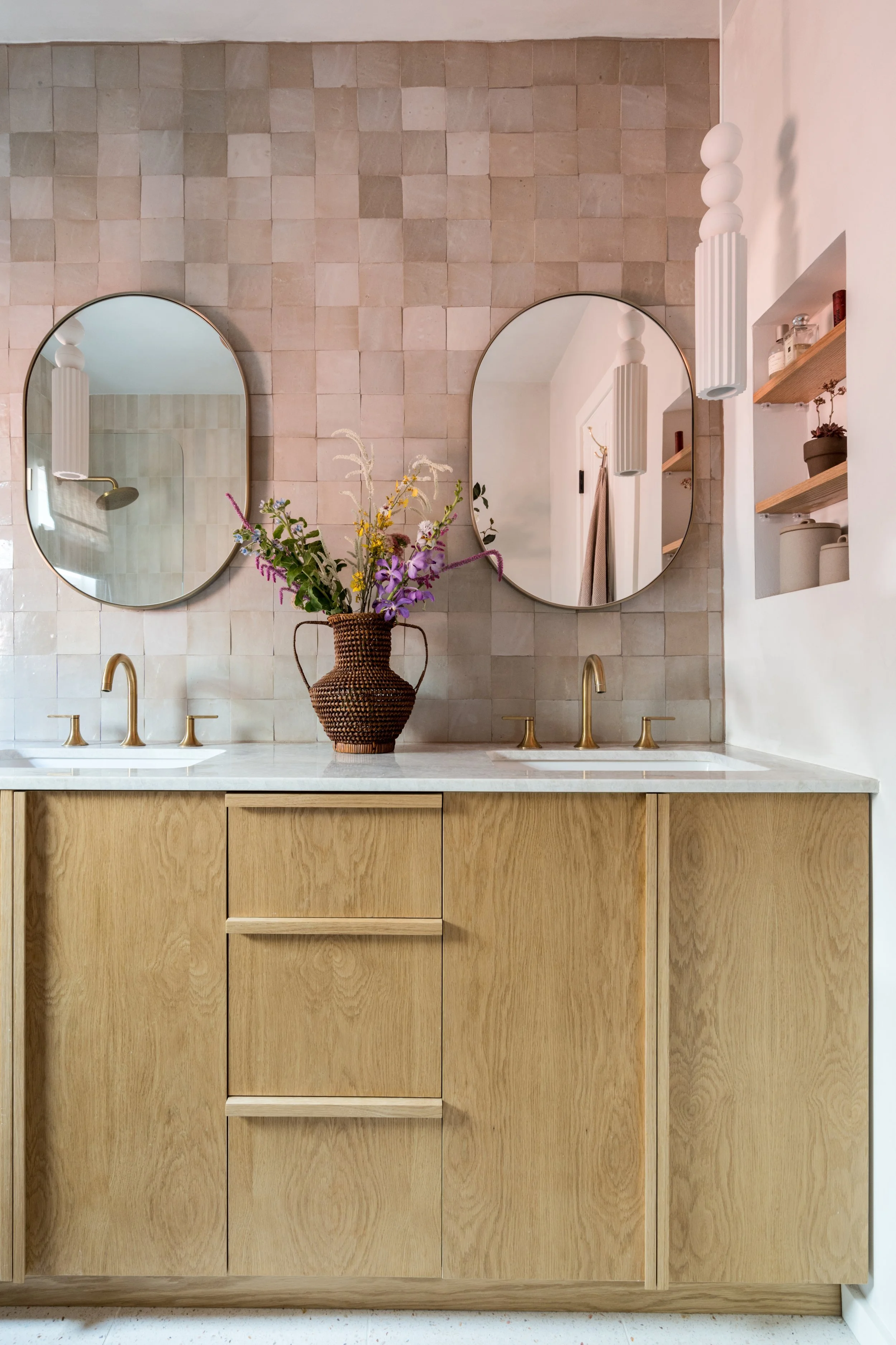 A bathroom vanity with a wooden cabinet, dual sinks with brass faucets, two oval mirrors, a decorative vase with flowers, and wall-mounted pendant lights. There are plants and toiletries on open shelves.