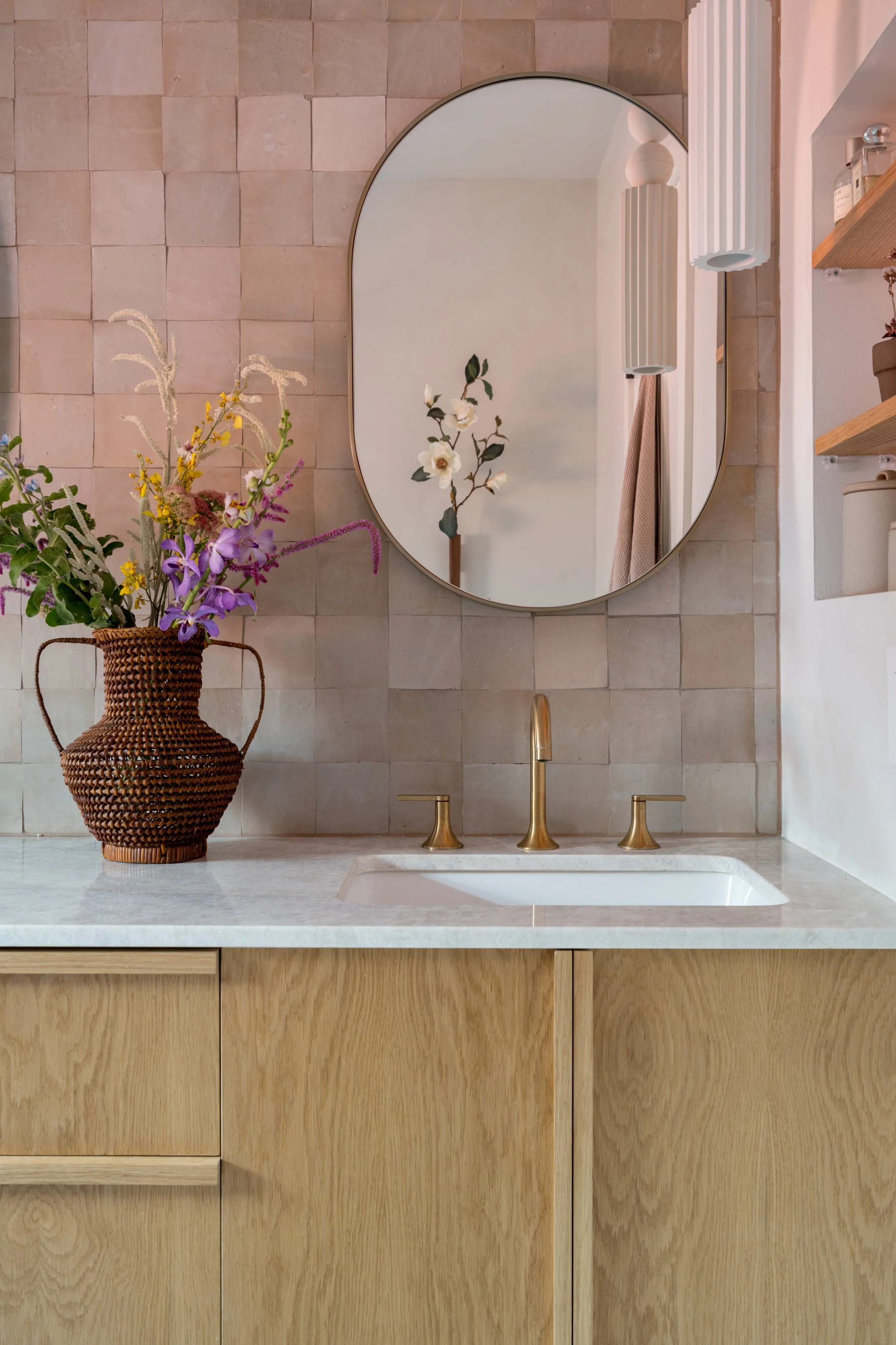 A bathroom vanity with a marble countertop, brass fixtures, a wicker vase with flowers, a large oval mirror, and open shelves with toiletries and decor, set against a beige tiled wall.