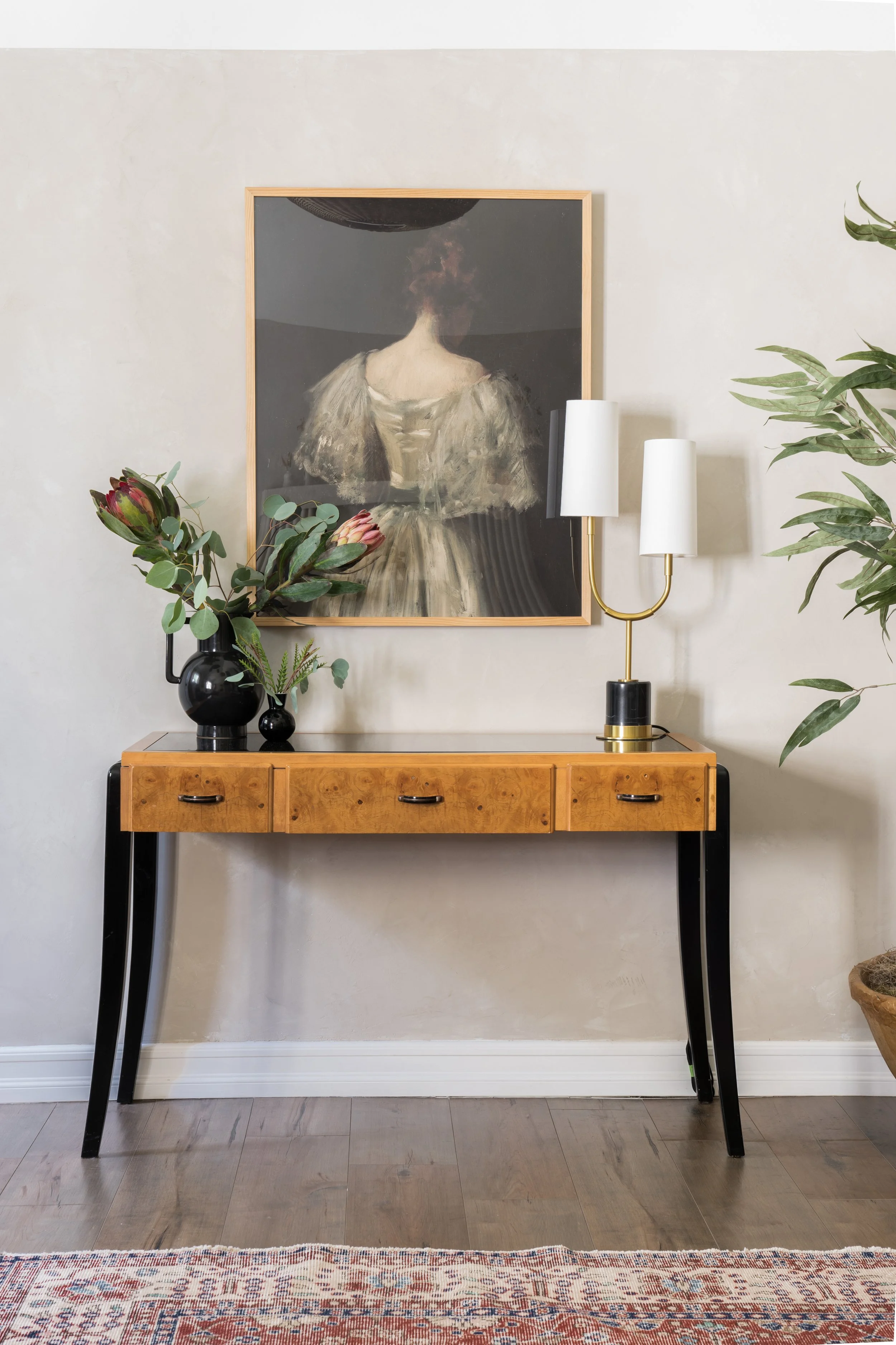 A wooden console table with black legs against a beige wall. On the table, there's a black vase with pink protea flowers and greenery, and a black smaller vase. A modern gold and white lamp with two white shades is also on the table. A framed painting of a woman with her back turned, wearing a dress with fur sleeves, hangs on the wall above. To the right, part of a large leafy green plant is visible.