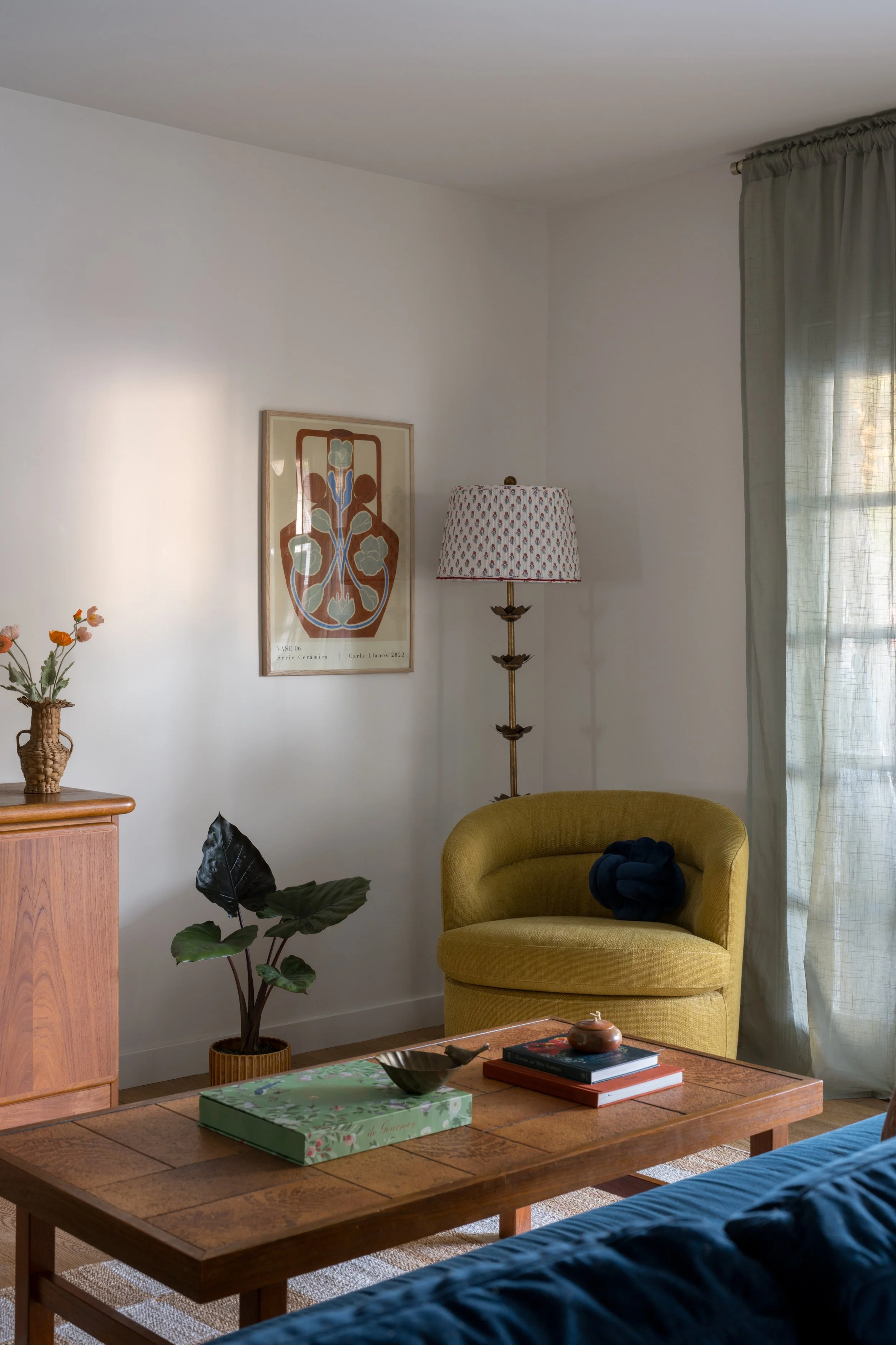 Living room with a yellow armchair, a tall lamp with a patterned shade, a wooden coffee table with books, a green plant in a basket, and a framed art piece on a white wall near a window with curtains.