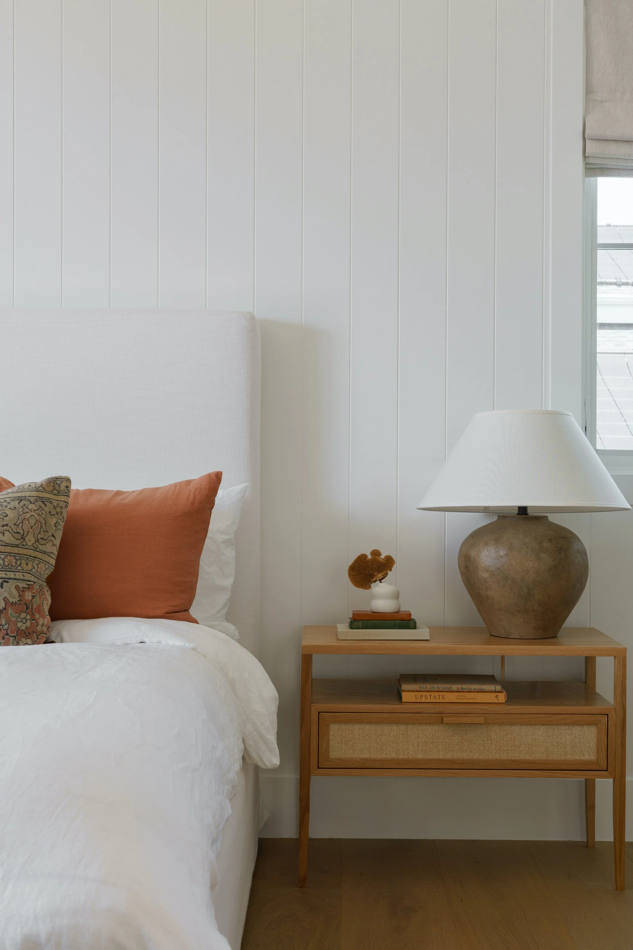 A bedroom corner with a white bed and a wooden nightstand holding a large clay-colored lamp, a stack of books, and decorative items.