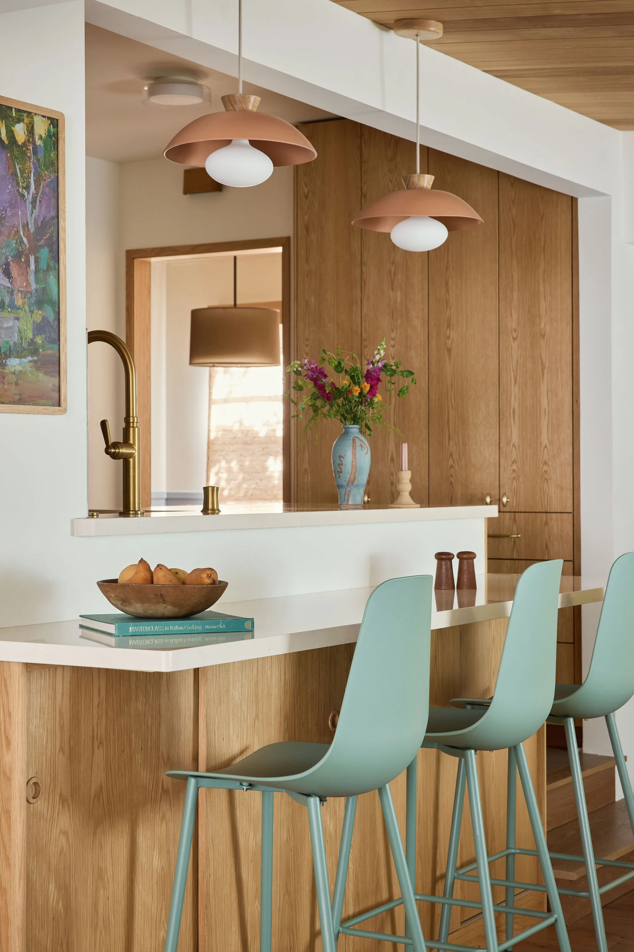 Modern kitchen interior with a wooden island, teal bar stools, pendant lights, a vase with flowers, and a brass faucet.