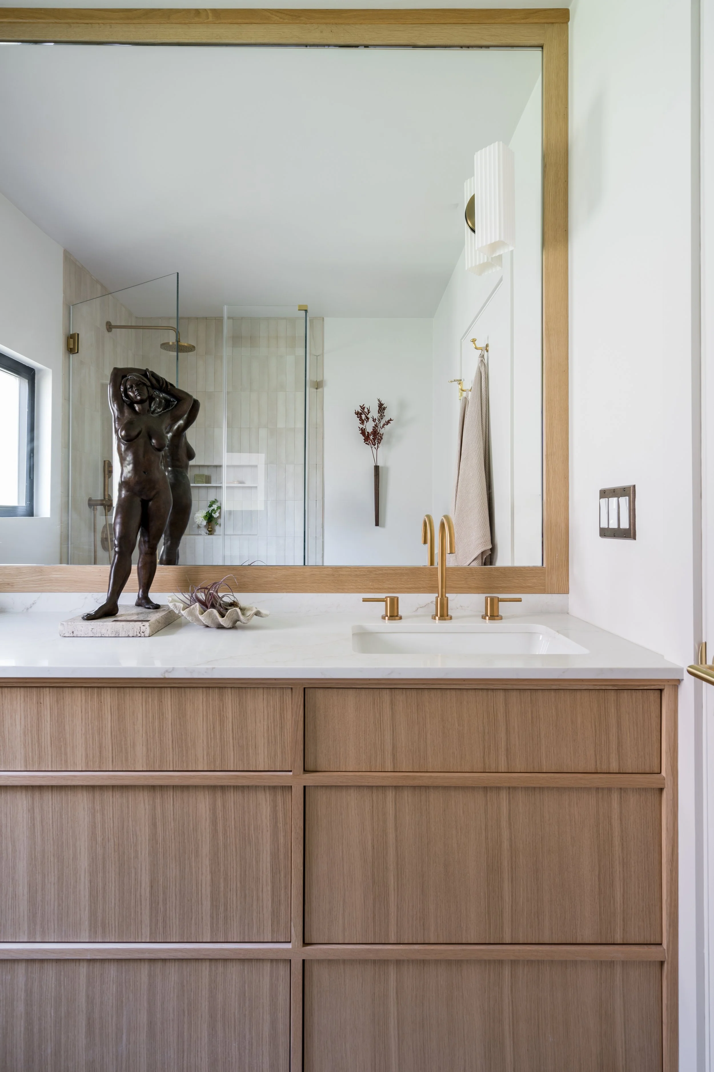 Modern bathroom vanity with a large mirror, a decorative bronze sculpture of a woman, and a view into the shower area with a glass door and built-in shelves, in neutral tones with brass fixtures.