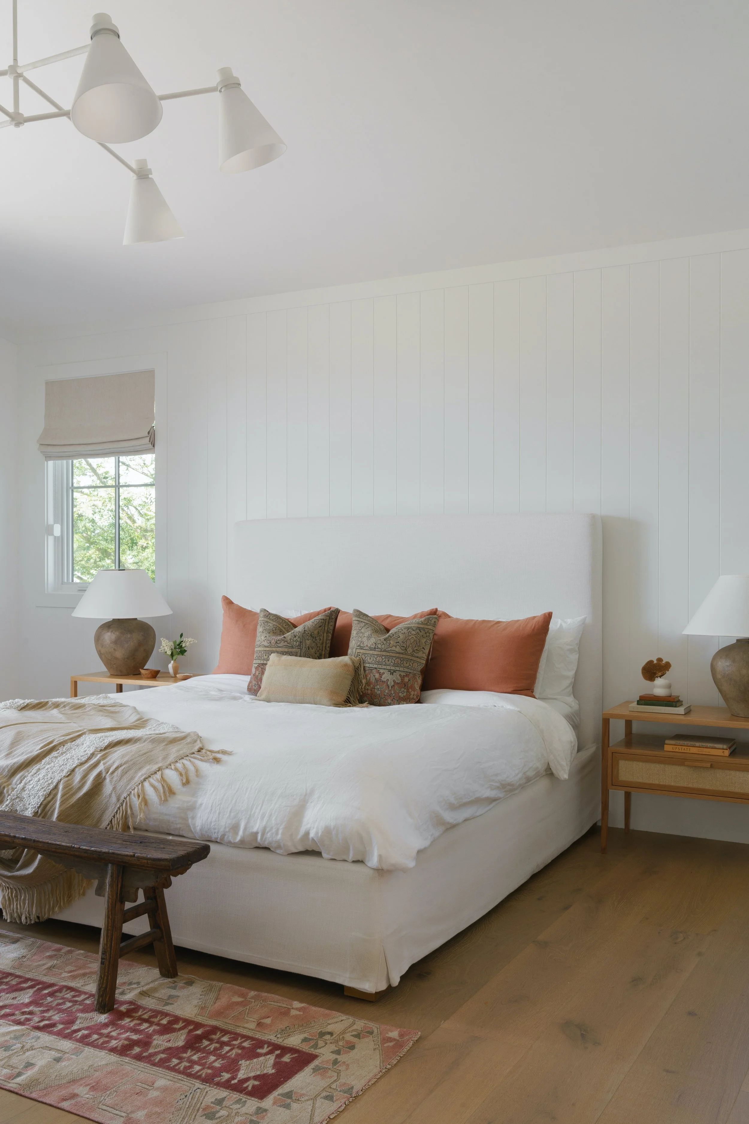 A cozy bedroom featuring a white upholstered bed with rust-colored and patterned pillows, wooden nightstands with lamps, a window with a beige Roman shade, a wooden bench at the foot of the bed, and a patterned area rug, all in a bright white-walled room.