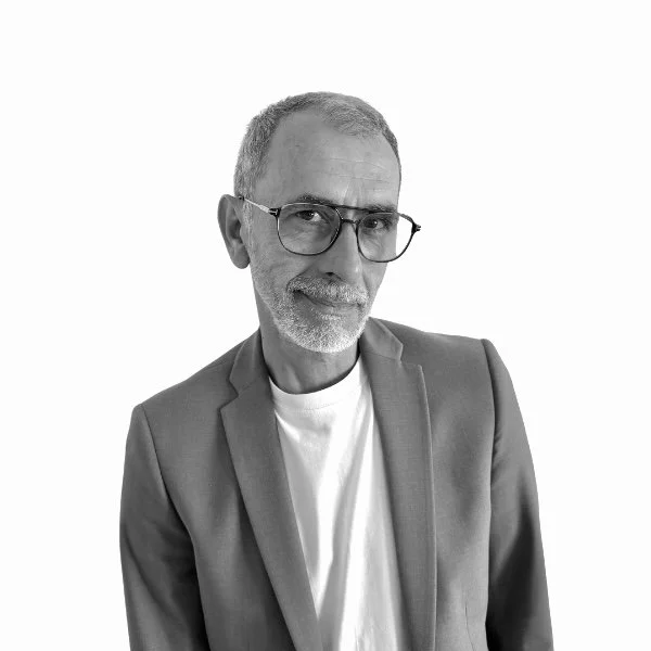Portrait en noir et blanc d'un homme moustachu portant des lunettes, une veste et un t-shirt blanc, regardant vers la caméra avec un léger sourire.