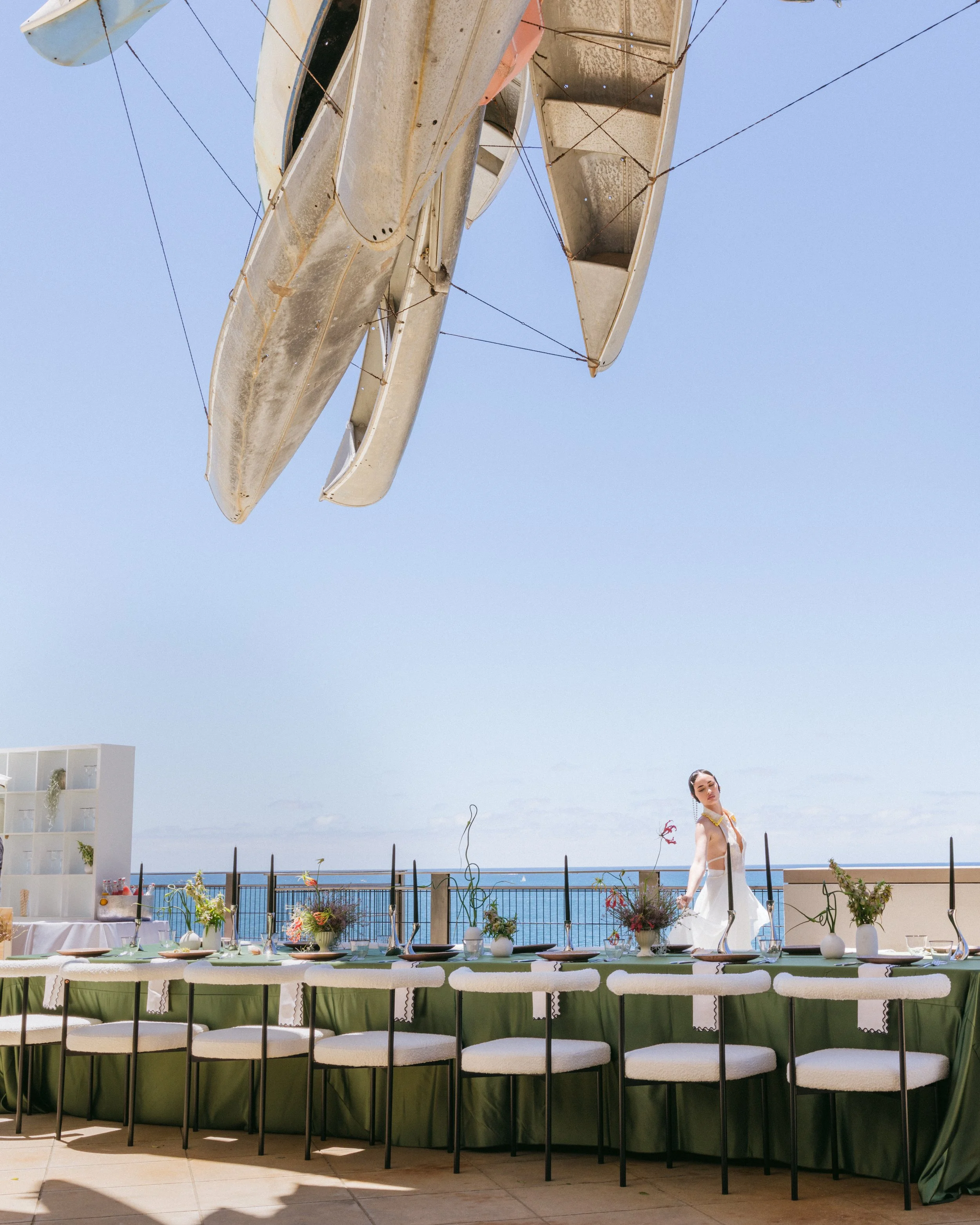 Outdoor oceanfront wedding reception with green linen banquet table, bouclé dining chairs, colorful floral arrangements, and dark taper candles on a rooftop terrace beneath a large suspended sculpture against a clear blue sky.