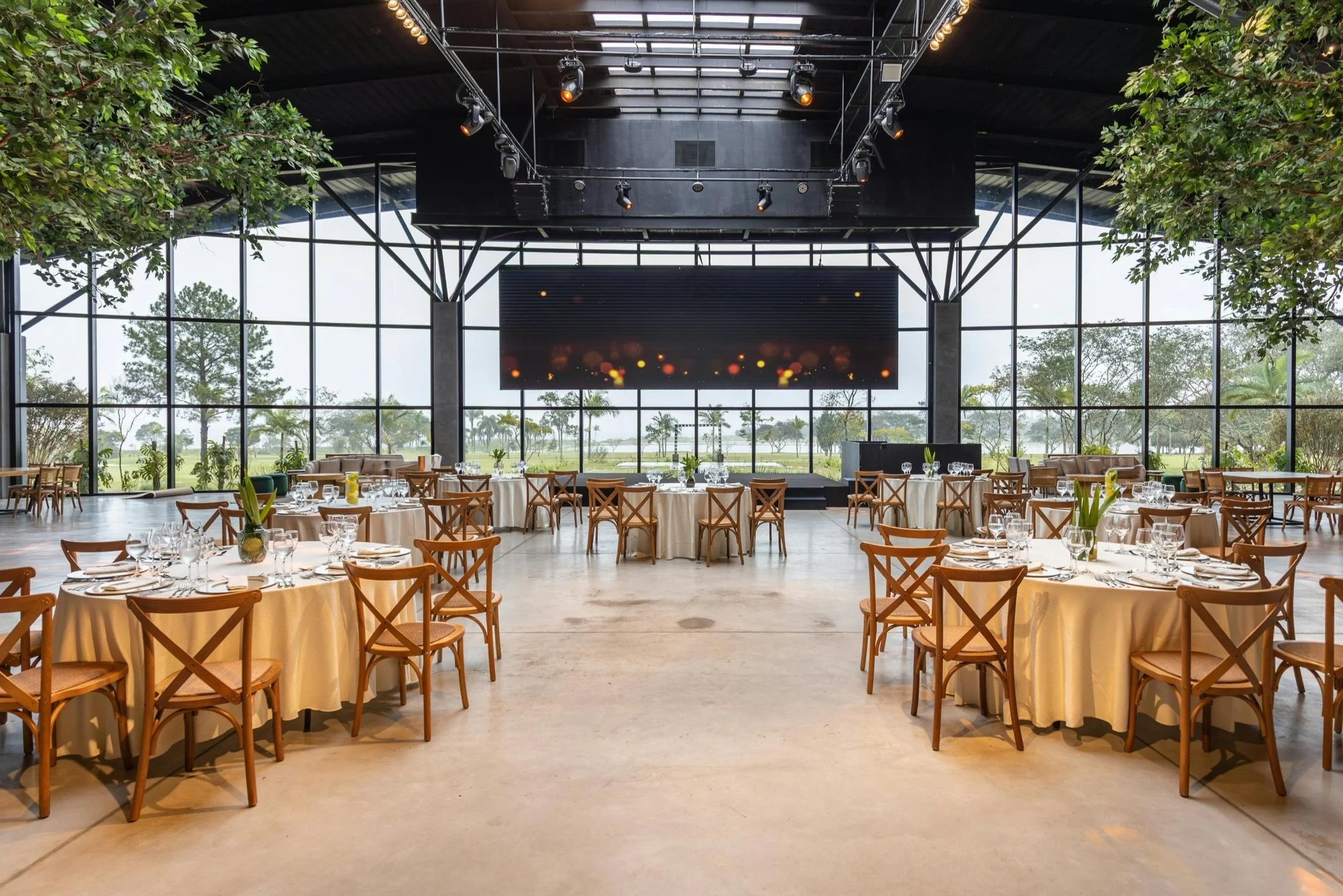 Event space with round tables covered with white tablecloths, set with glassware and cutlery, inside a large glass-walled building with greenery outside, pendant lighting on ceiling, and a large black stage with a digital screen in the background.