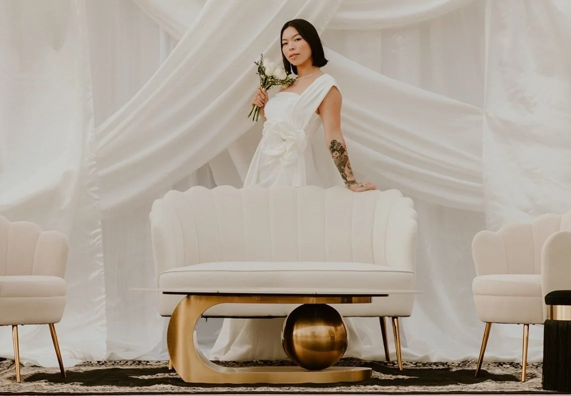 A woman in a white dress holding a bouquet of flowers, standing behind a white sofa with gold accents in a room decorated with white drapes and furniture.