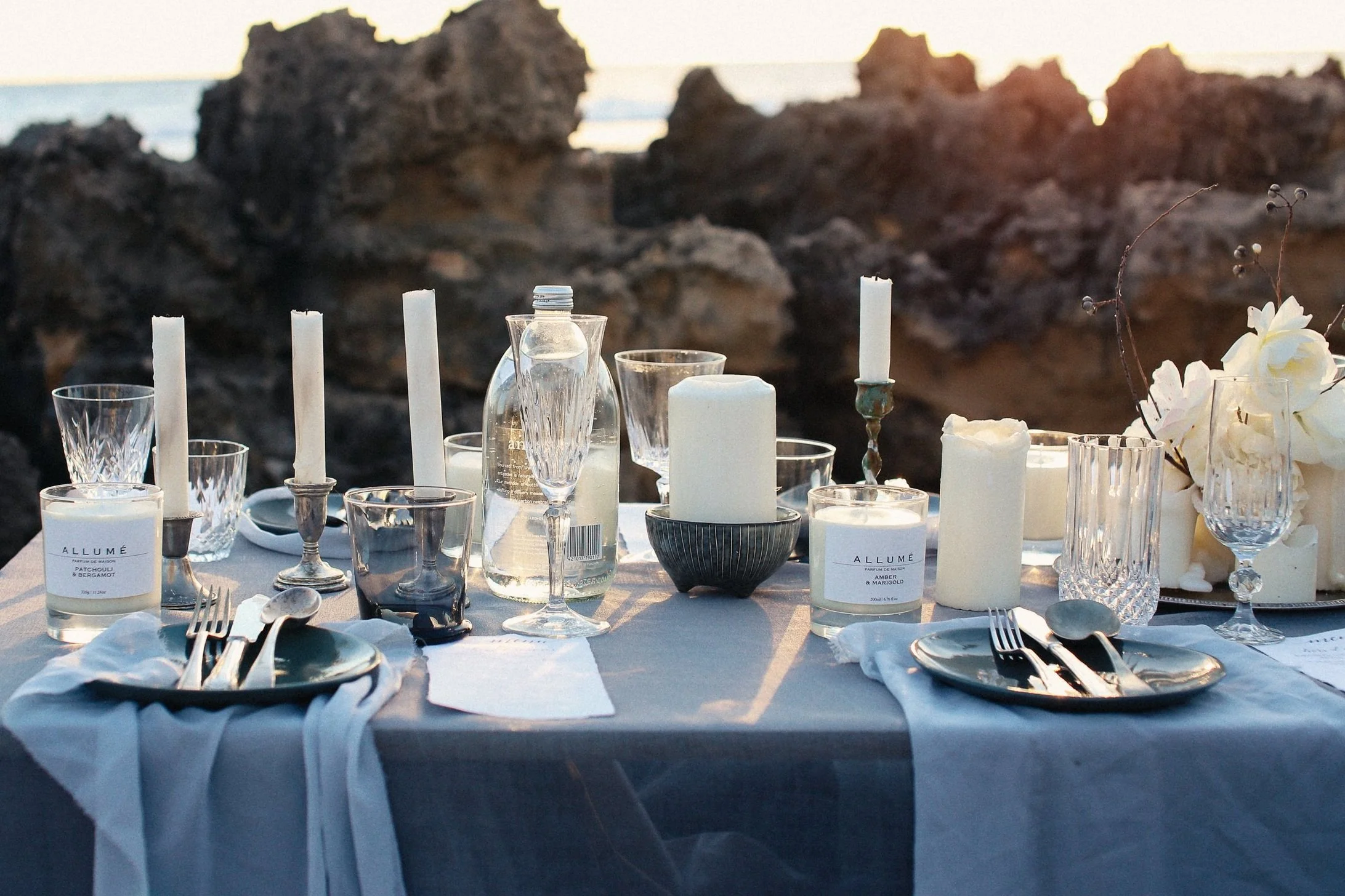 Elegant outdoor dining table set with candles, glassware, plates, and silverware, overlooking rocks and the ocean at sunset.