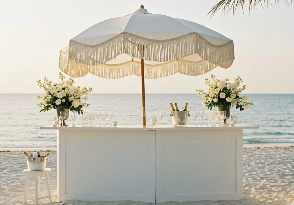 Beachside bar setup with white flowers, glasses, and champagne bottles under a large white parasol, near the ocean.