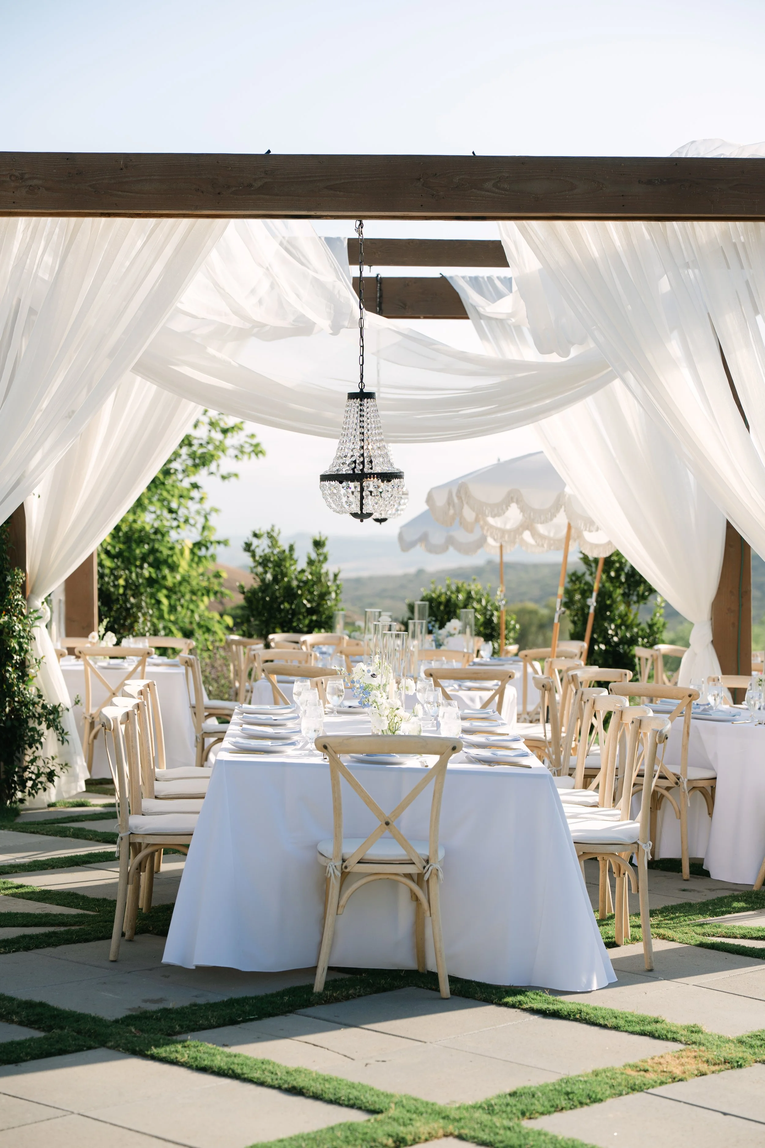 Elegant outdoor wedding reception setup with long rectangular tables, white tablecloths, wooden chairs, glassware, and floral centerpieces, under a draped canopy with a chandelier and parasols, with a scenic landscape in the background.