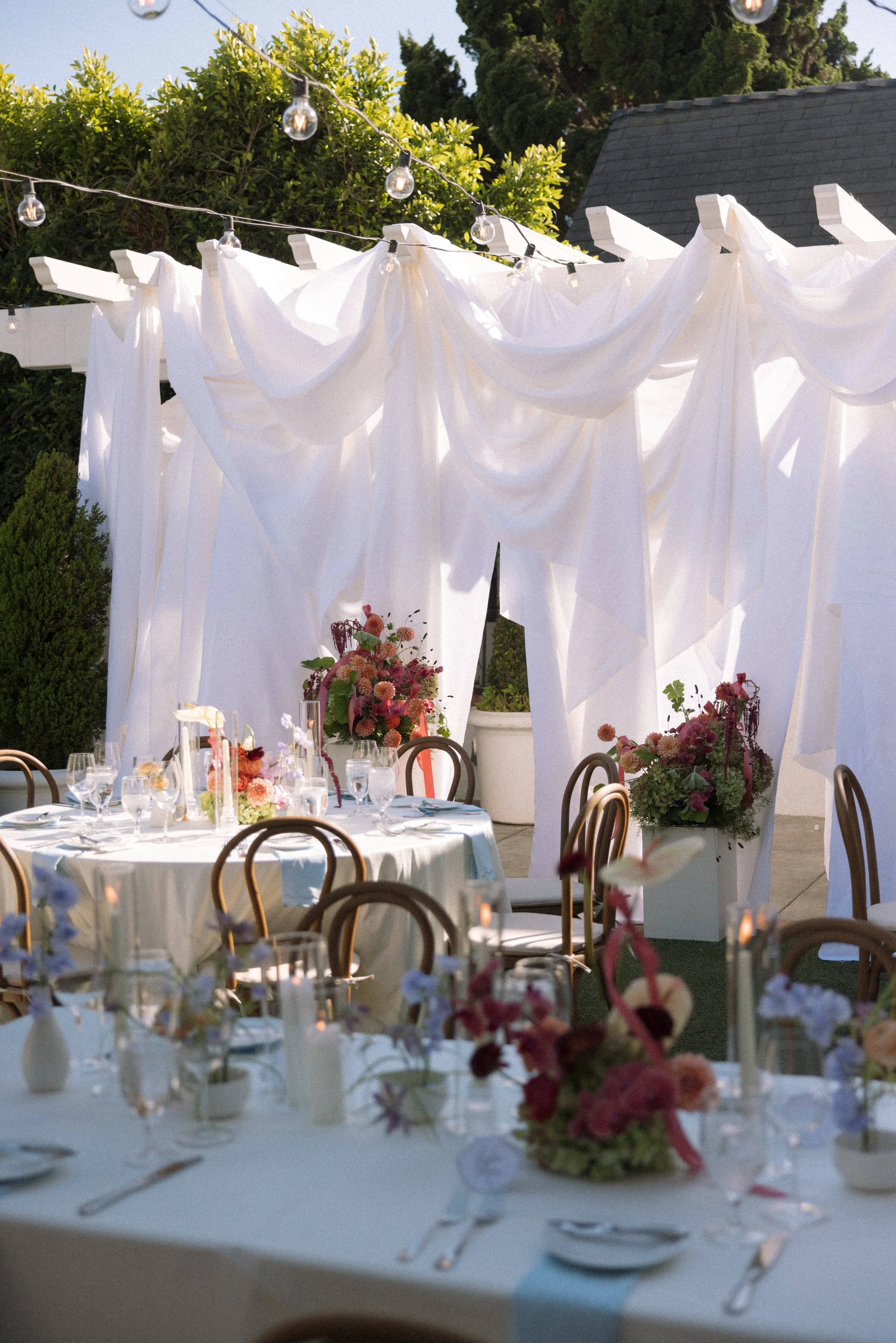 An outdoor wedding reception with round tables decorated with floral centerpieces, white tablecloths, and glassware. White drapery panels and string lights are set up overhead, with greenery in the background.