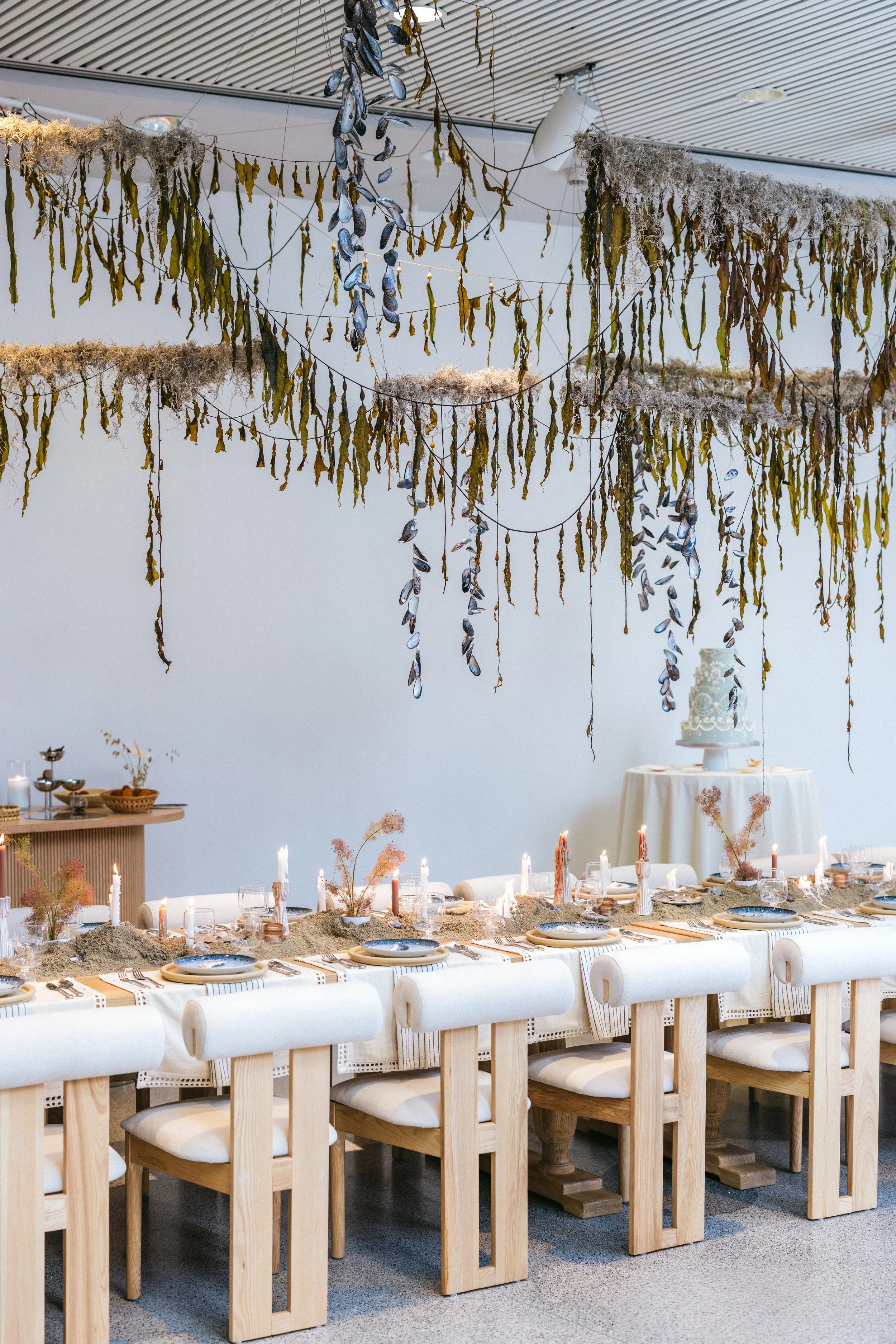Long banquet table set with sand-toned place settings, wooden chargers, blue accent plates, and tapered candles in terracotta holders, surrounded by cream upholstered dining chairs with natural wood frames.