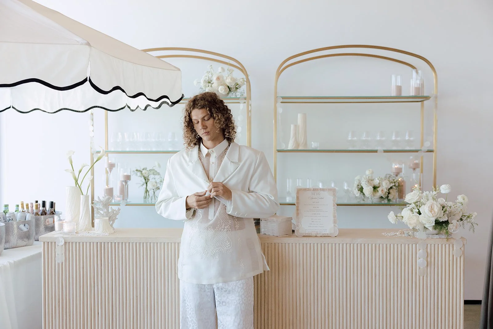 Fluted wood bar with gold-framed arched back shelves styled with glassware, candles, and white florals at a luxury wedding, accented by a black-and-white scalloped awning and signature cocktail menu sign.