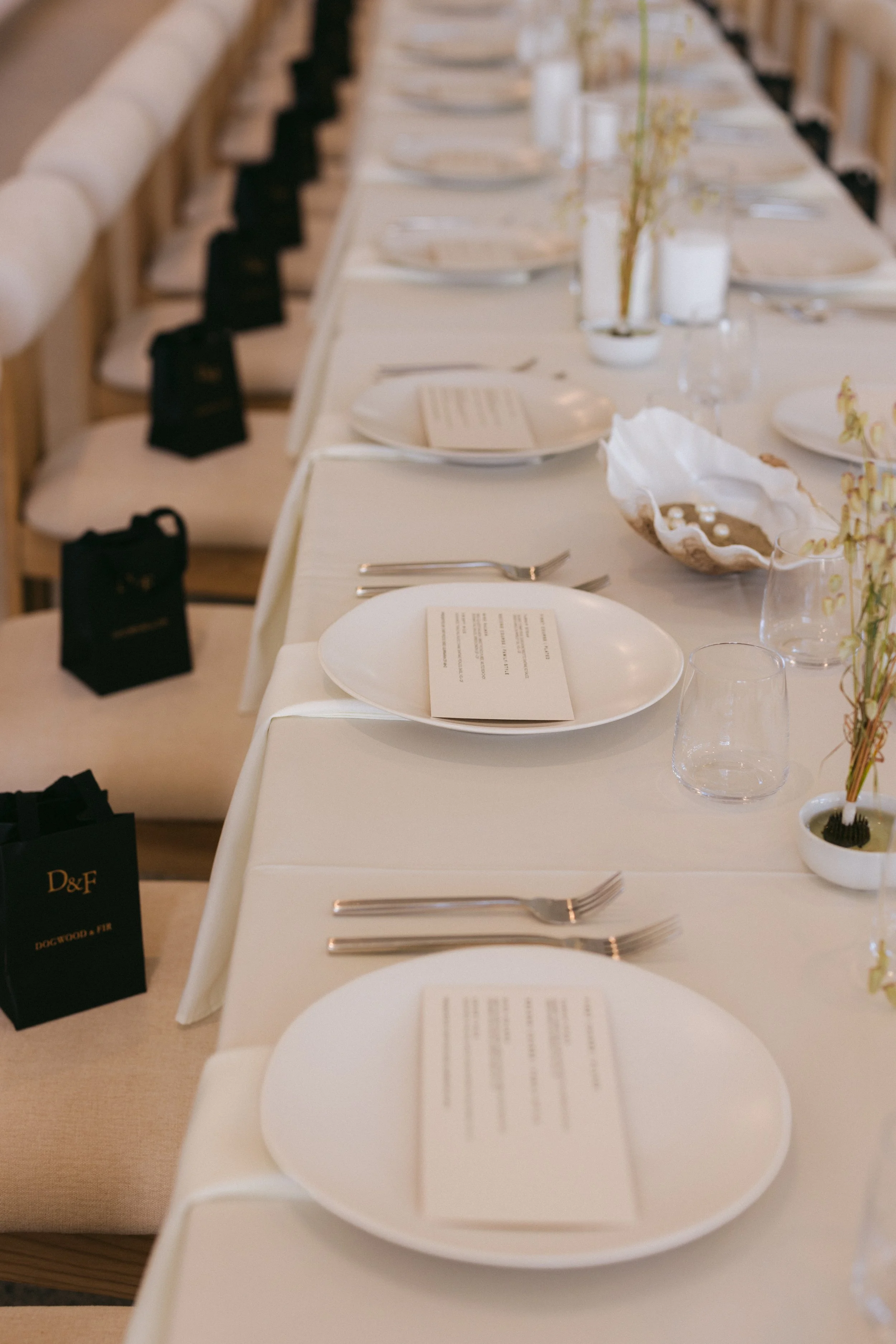 Long banquet table set with white plates, printed menus, silver flatware, stemless glasses, shell accents, and ceramic bud vases. Cream upholstered chairs with wood frames line the table, with black Dogwood & Fir gift bags at each seat.