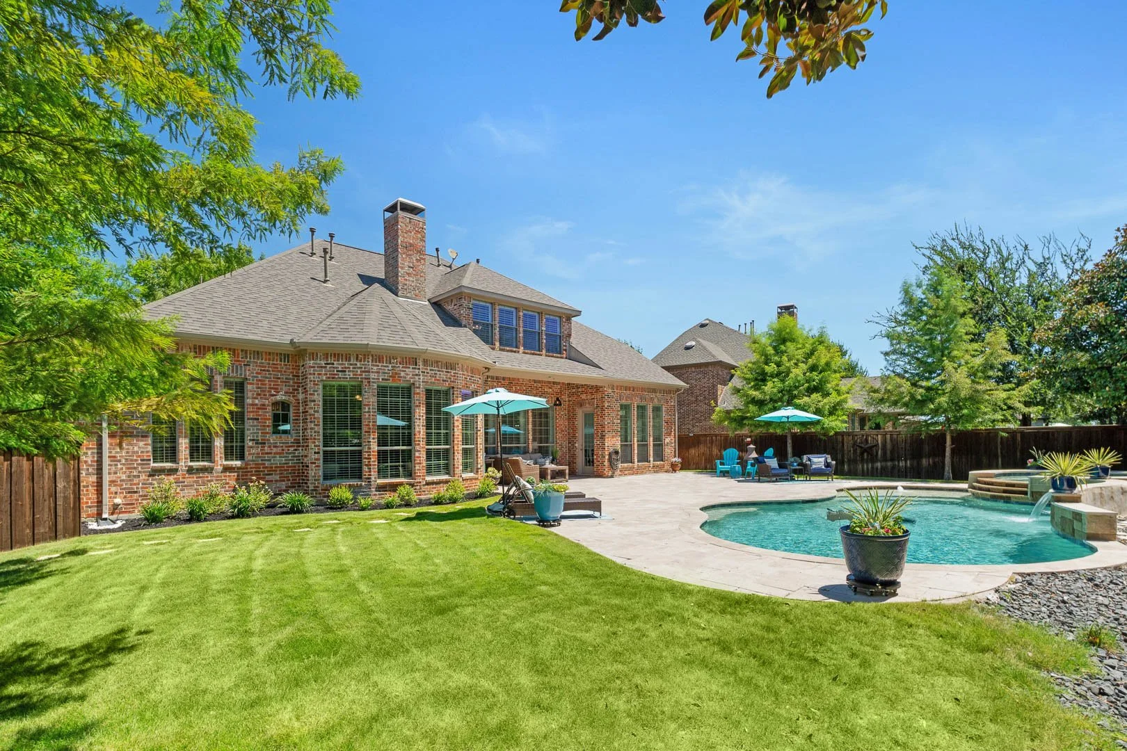Backyard view with a brick house, swimming pool, green lawn, patio area with umbrellas and outdoor furniture, surrounded by trees and a wooden fence, under a clear blue sky.